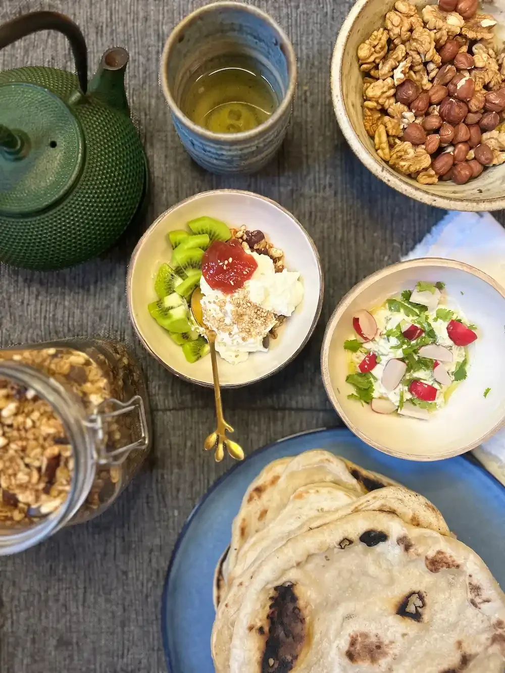 A breakfast table with a teapot, a glass of green tea, a bowl of mixed nuts, a bowl of fresh fruit with yogurt and granola, a bowl of salad with radishes and greens, and a plate of flatbreads on a rustic wooden surface.