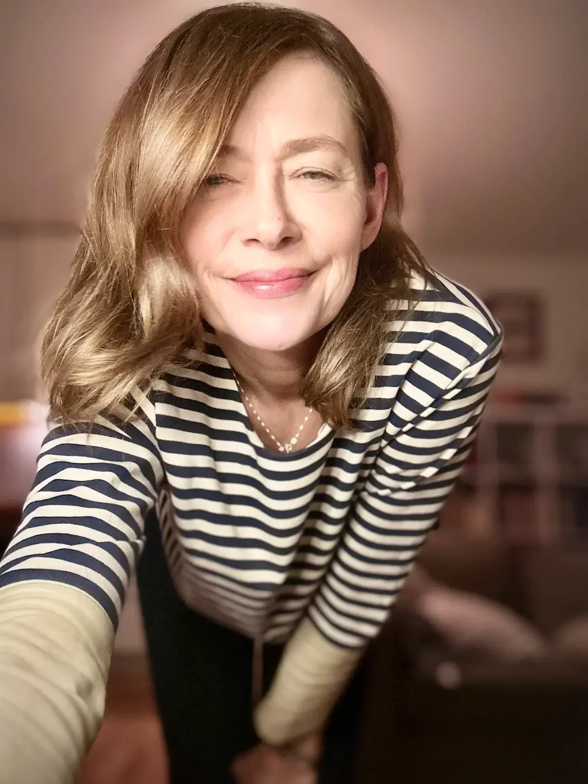 A woman with shoulder-length wavy hair, smiling and winking, wearing a black and white striped shirt and a pearl necklace in a cozy indoor setting.