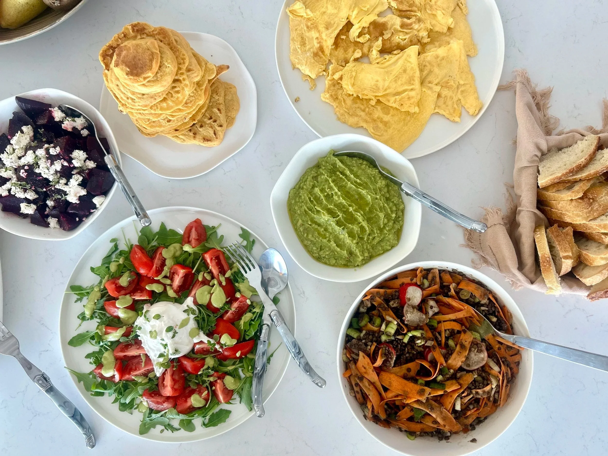 Assorted dishes on a white table including a beet salad topped with cheese, a tomato and lettuce salad with greens, a bowl of guacamole, black lentil and vegetable salad, a bowl of yellow scrambled eggs, and a plate of toasted bread slices.