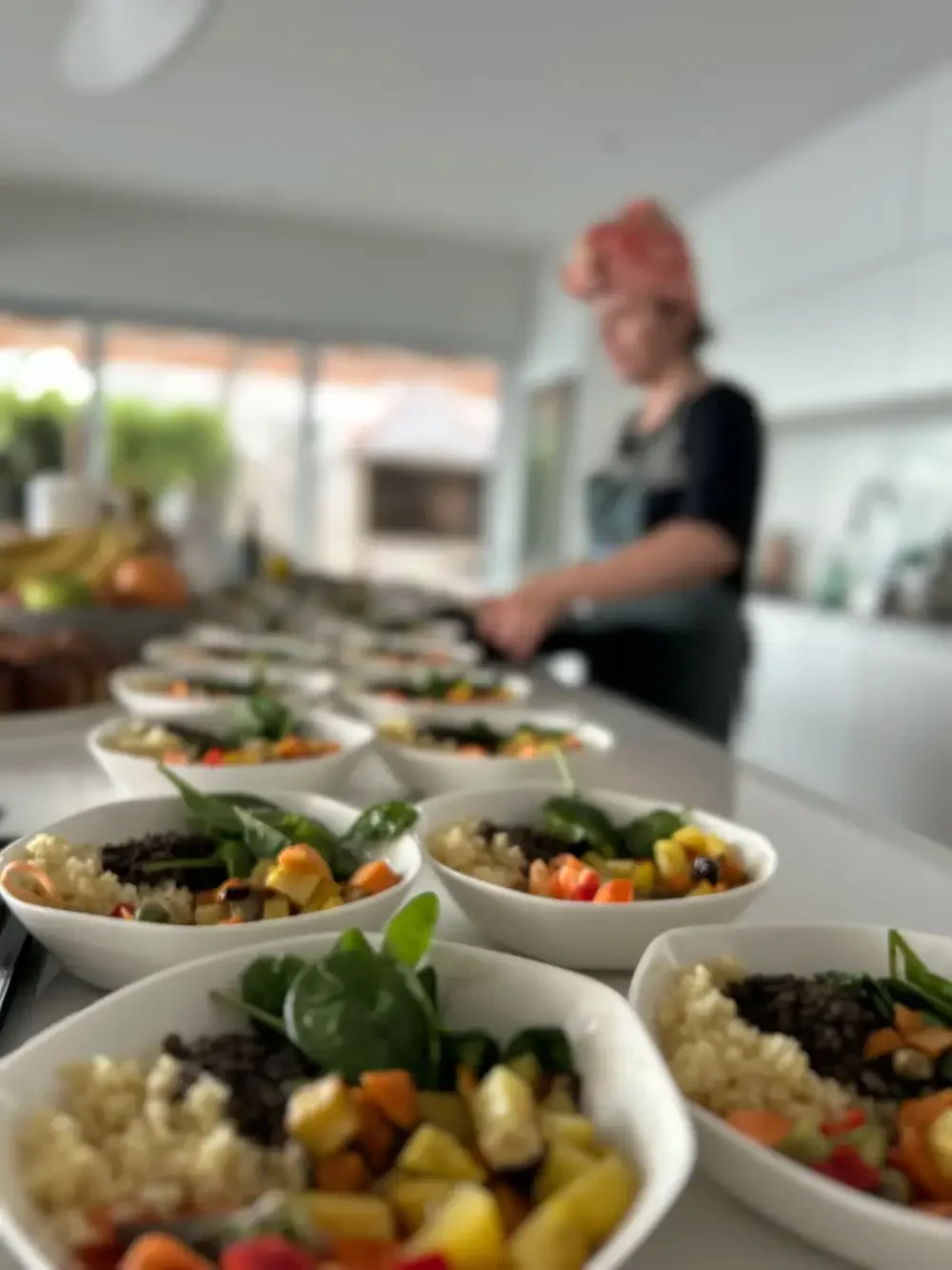Multiple bowls of colorful food, likely salads or grain bowls, on a kitchen counter with a woman in the background preparing more dishes.