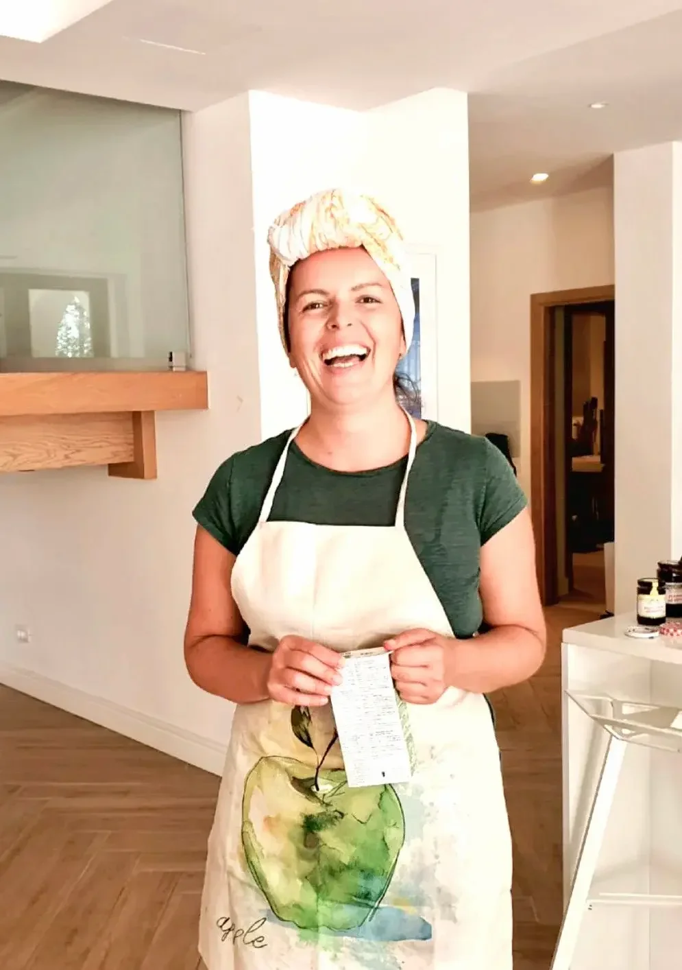 Woman smiling in a kitchen wearing a green shirt, a colorful headwrap, and a beige apron with an apple illustration.