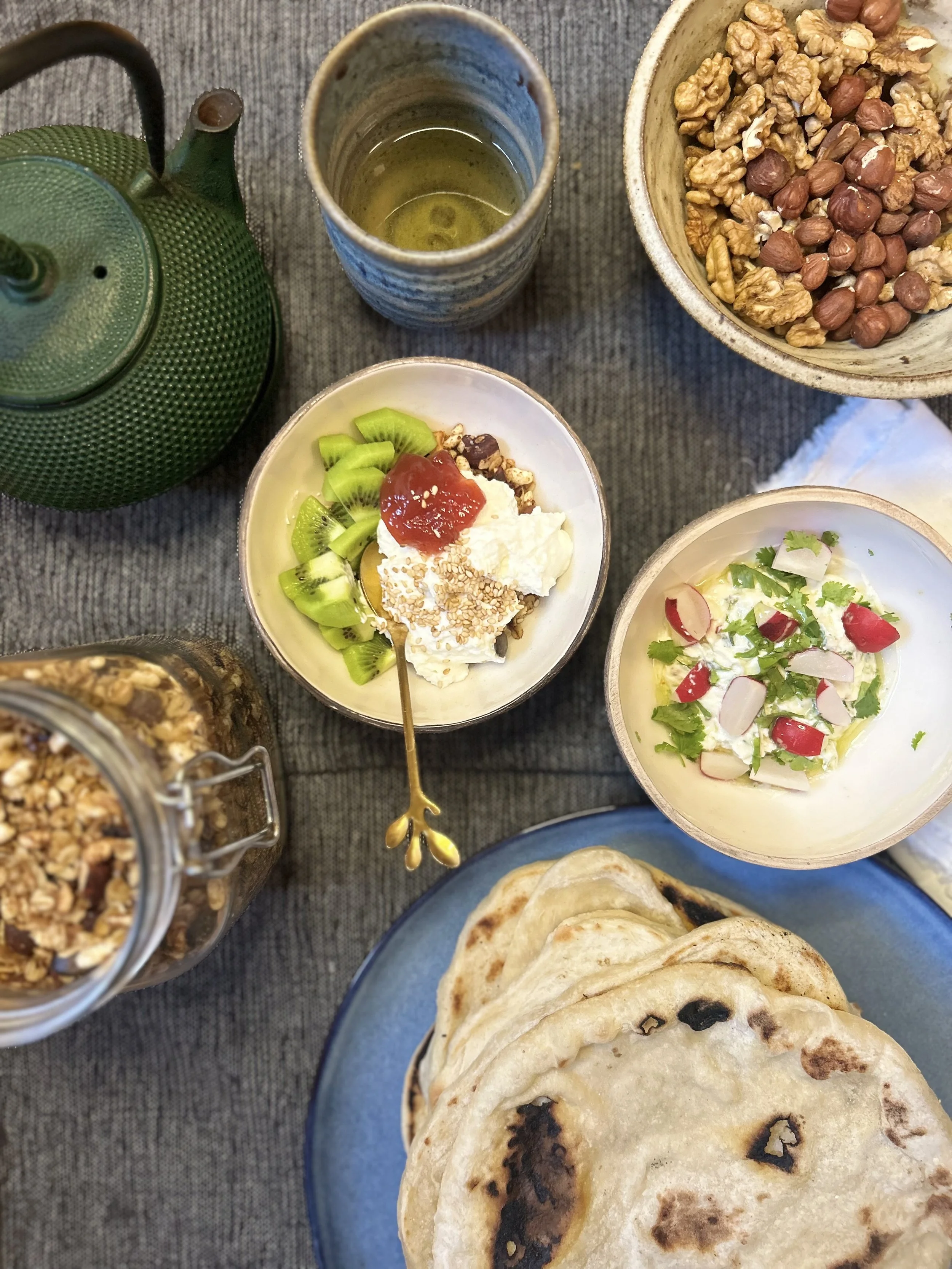 A top-down view of a breakfast spread including a plate of flatbread, bowls of yogurt with fruit and herbs, a jar of granola, a bowl of nuts, a glass of tea, and a small dish with yogurt, granola, kiwi, and toppings.