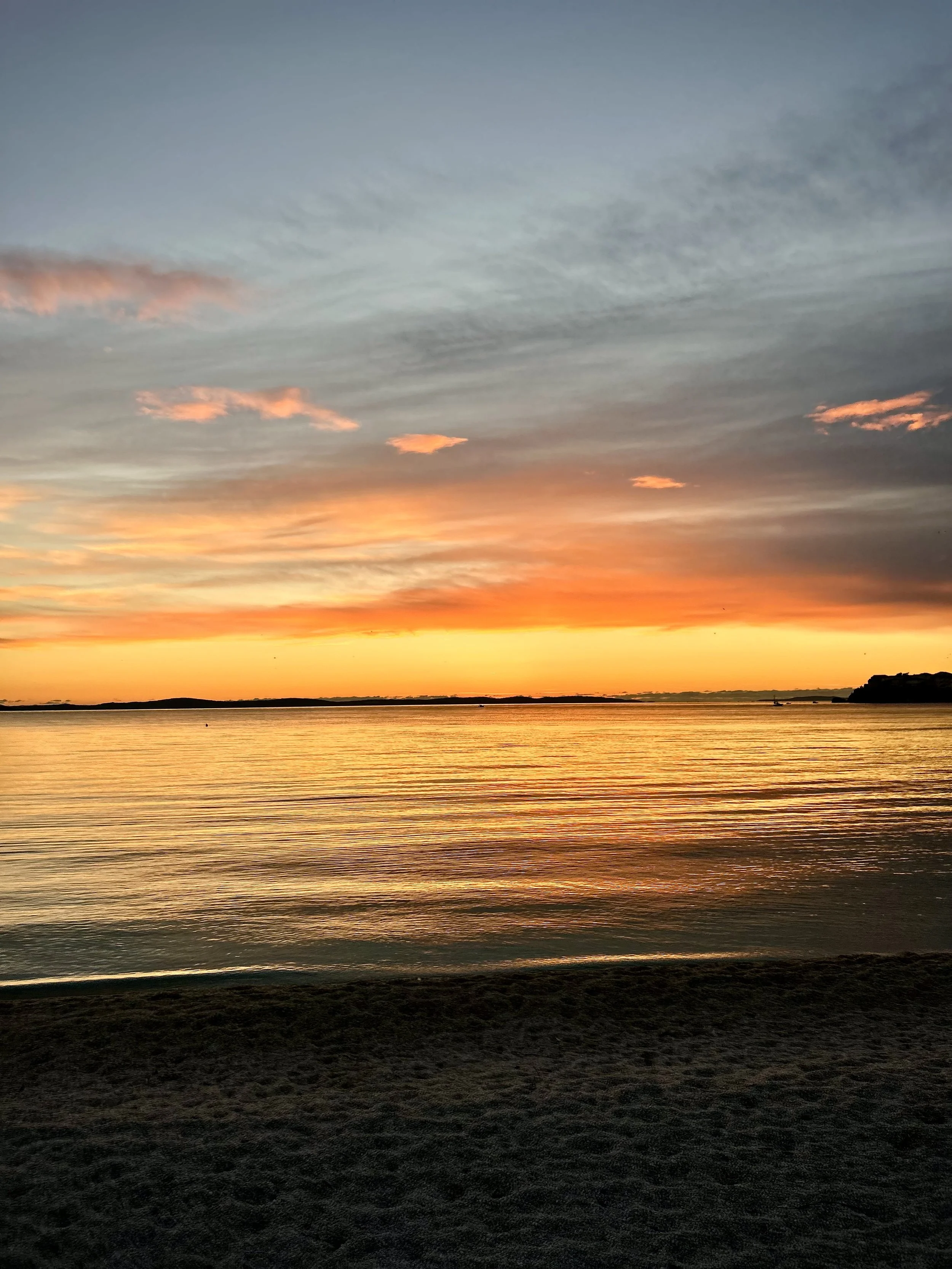 Sunset over a calm body of water with an orange and pink sky and a sandy beach in the foreground.