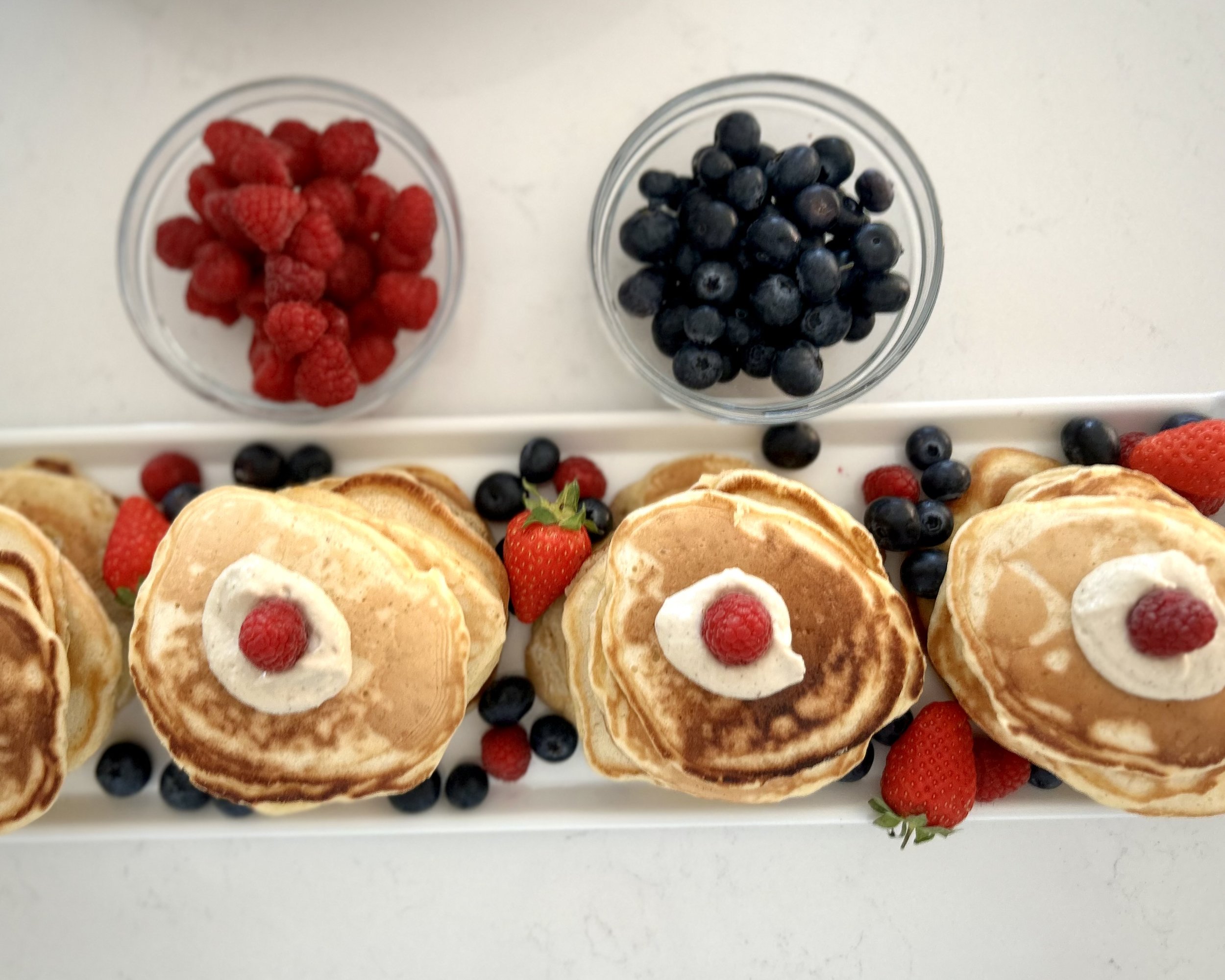 A plate of pancakes topped with whipped cream and raspberries, decorated with strawberries and blueberries, along with two bowls of raspberries and blueberries.