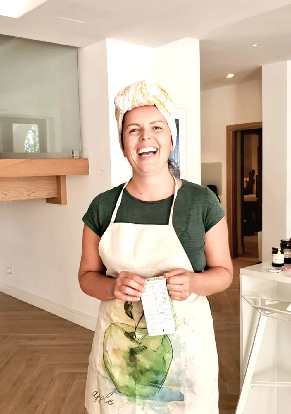 A woman smiling and laughing in a kitchen, wearing a beige apron with a green apple watercolor design and a beige headscarf, holding a small packet, with jars on the counter behind her.