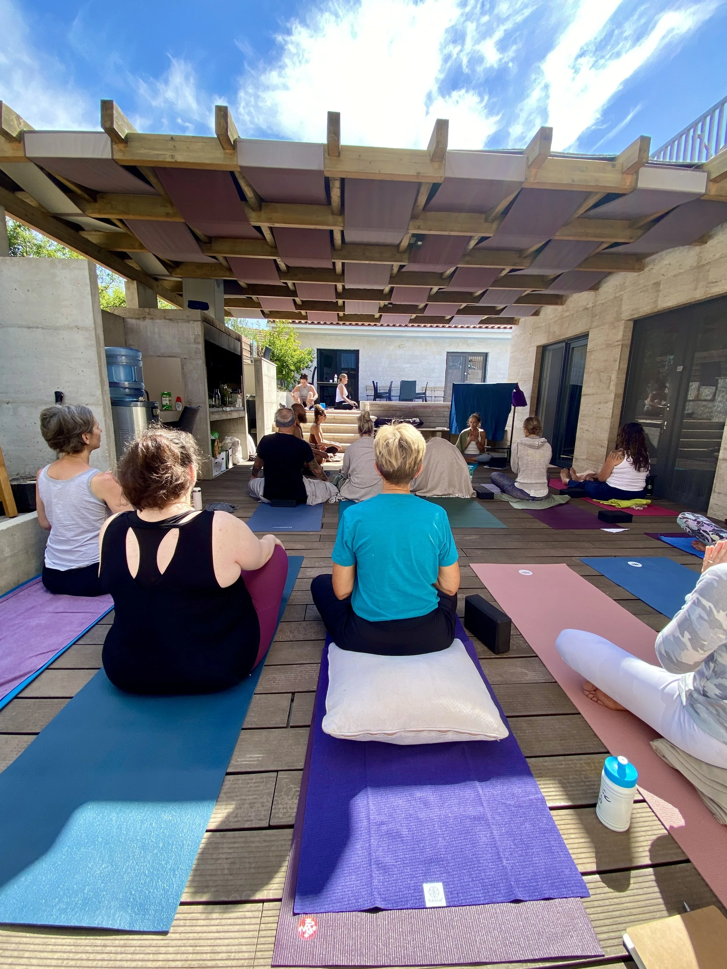 People practicing yoga outdoors on a wooden deck under a shaded wooden pergola, facing a stage area with instructors, with blue sky and clouds overhead.