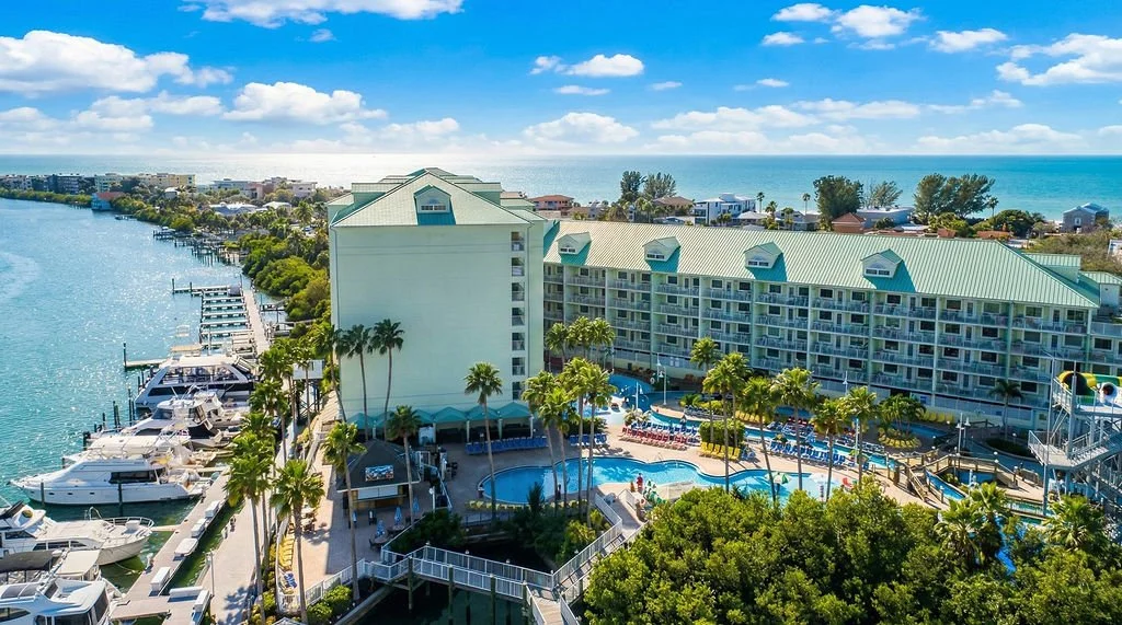 Aerial view of a beachfront resort with a swimming pool, palm trees, and boats docked along a canal, with the ocean in the background under a partly cloudy sky.