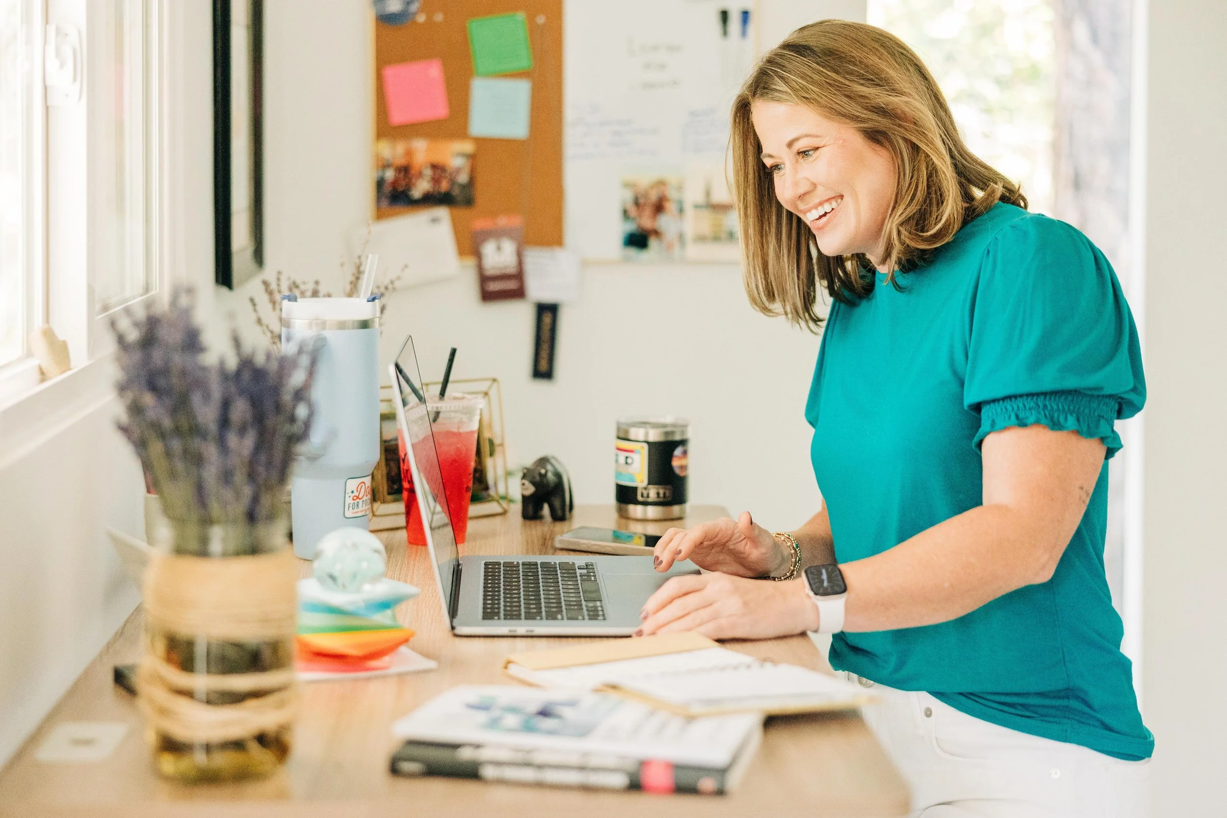 A woman in a teal shirt works at a cluttered desk with a laptop, notebooks, and colorful markers, smiling and holding her hands near the laptop keyboard.