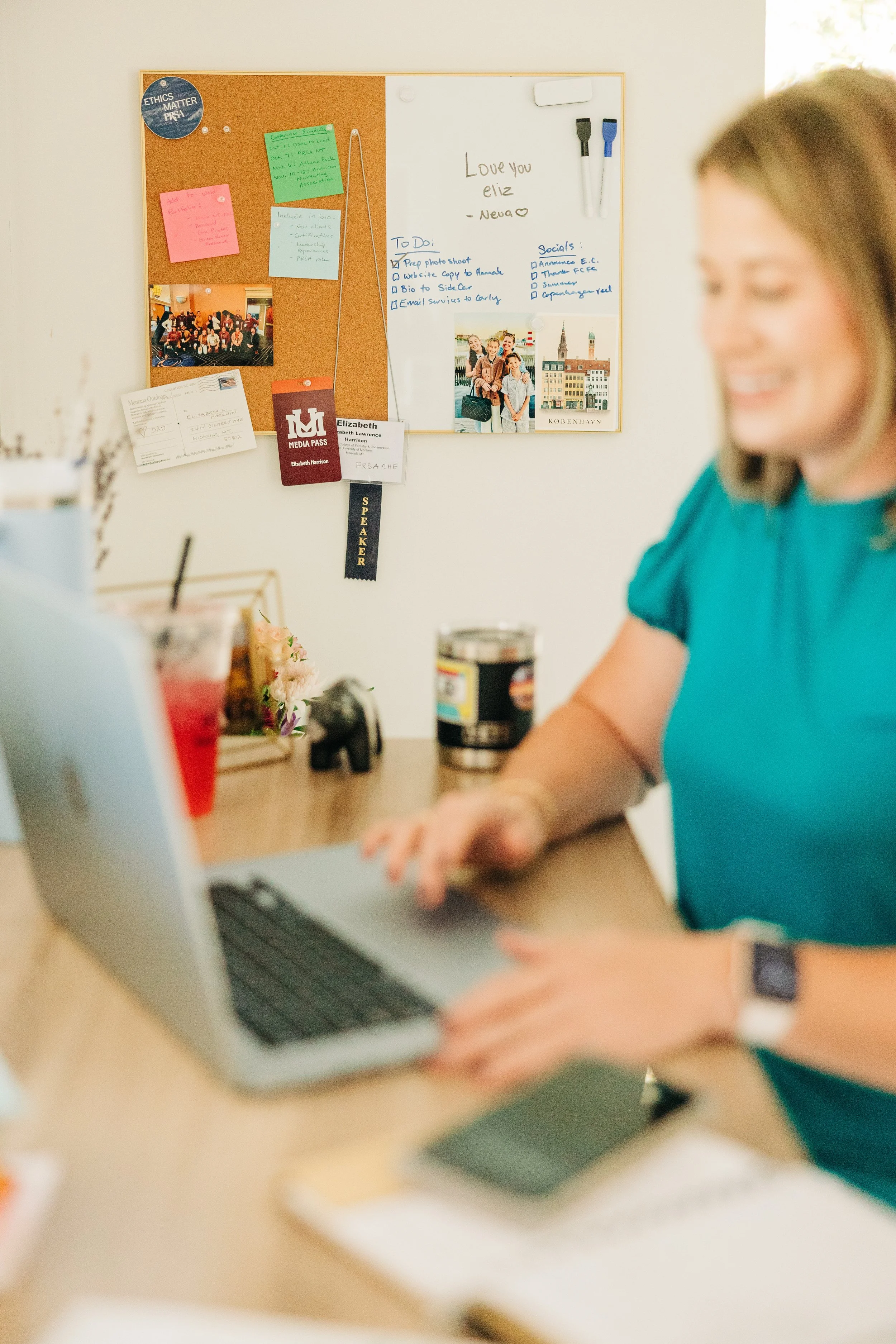 A woman sitting at a desk using a laptop with a notebook and phone in front of her. In the background, a whiteboard and corkboard with photos, notes, and a media pass hang on the wall.