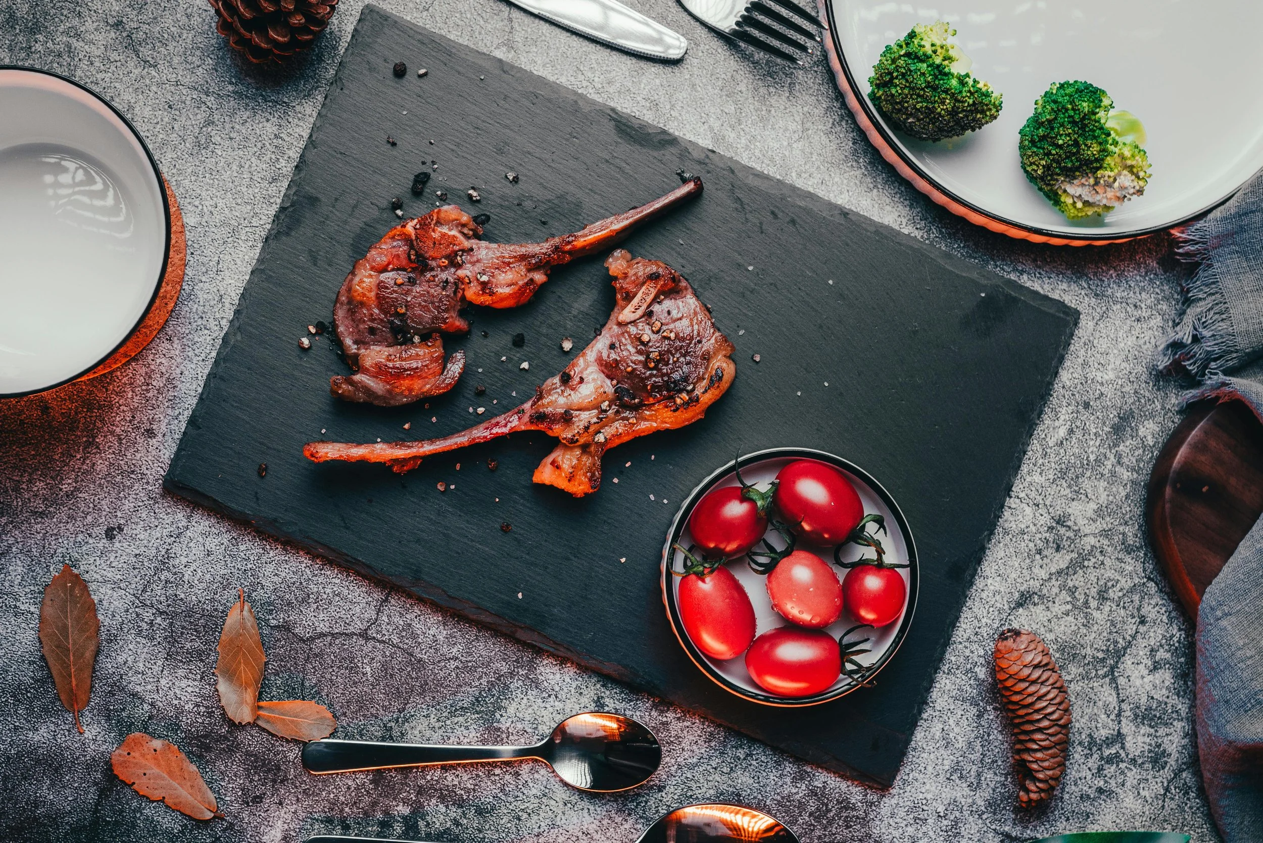 Cooked bacon strips on a black serving board, cherry tomatoes in a bowl, broccoli on a plate, a cup of water, and utensils on a table with a gray surface.