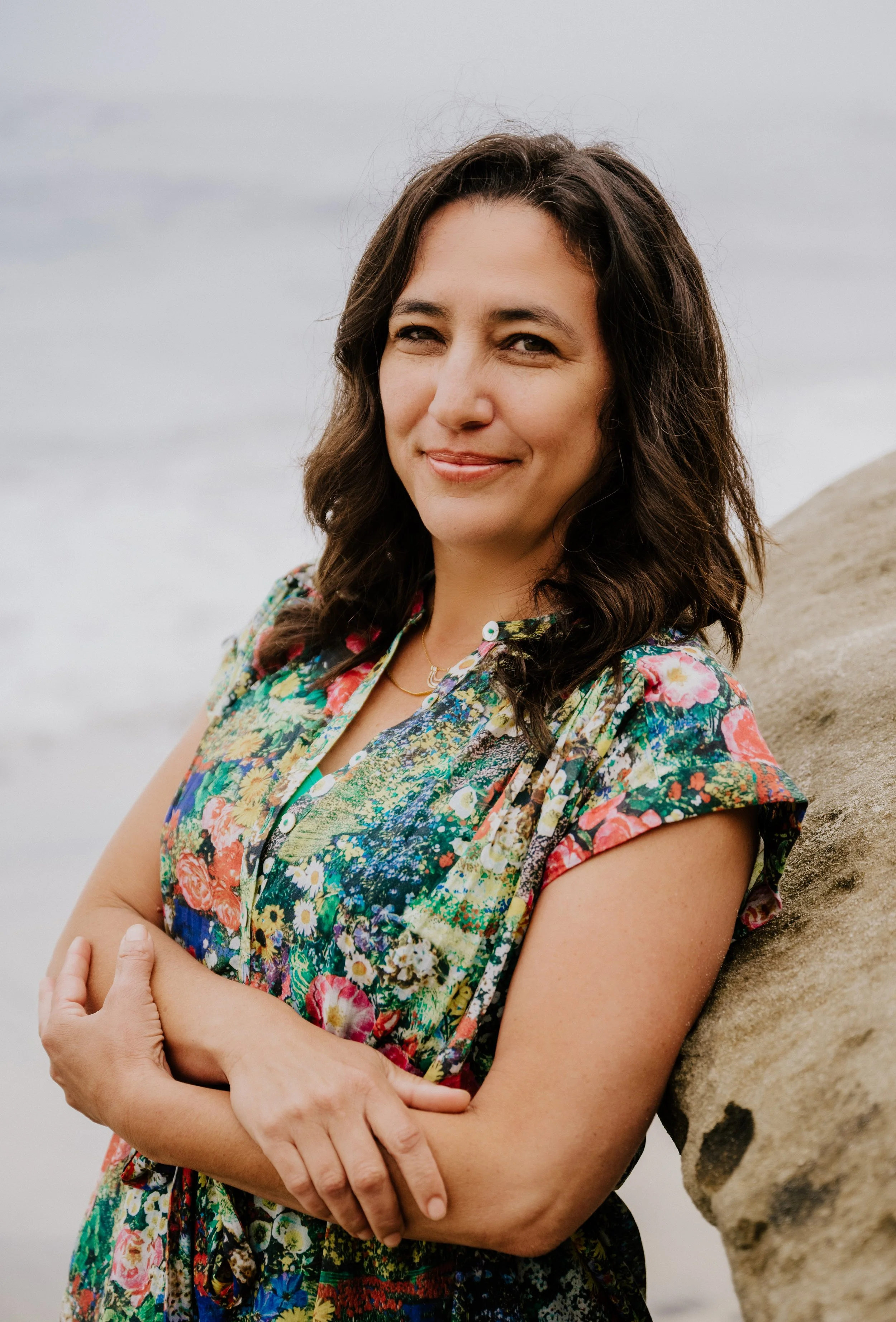 A woman with dark brown, curly hair, smiling and crossing her arms, standing next to a rock on a beach with cloudy sky in the background, wearing a colorful floral dress.