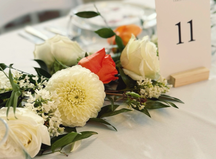 A floral centerpiece with white and orange roses, white dahlias, and greenery on a white tablecloth, with a table number 11 card.