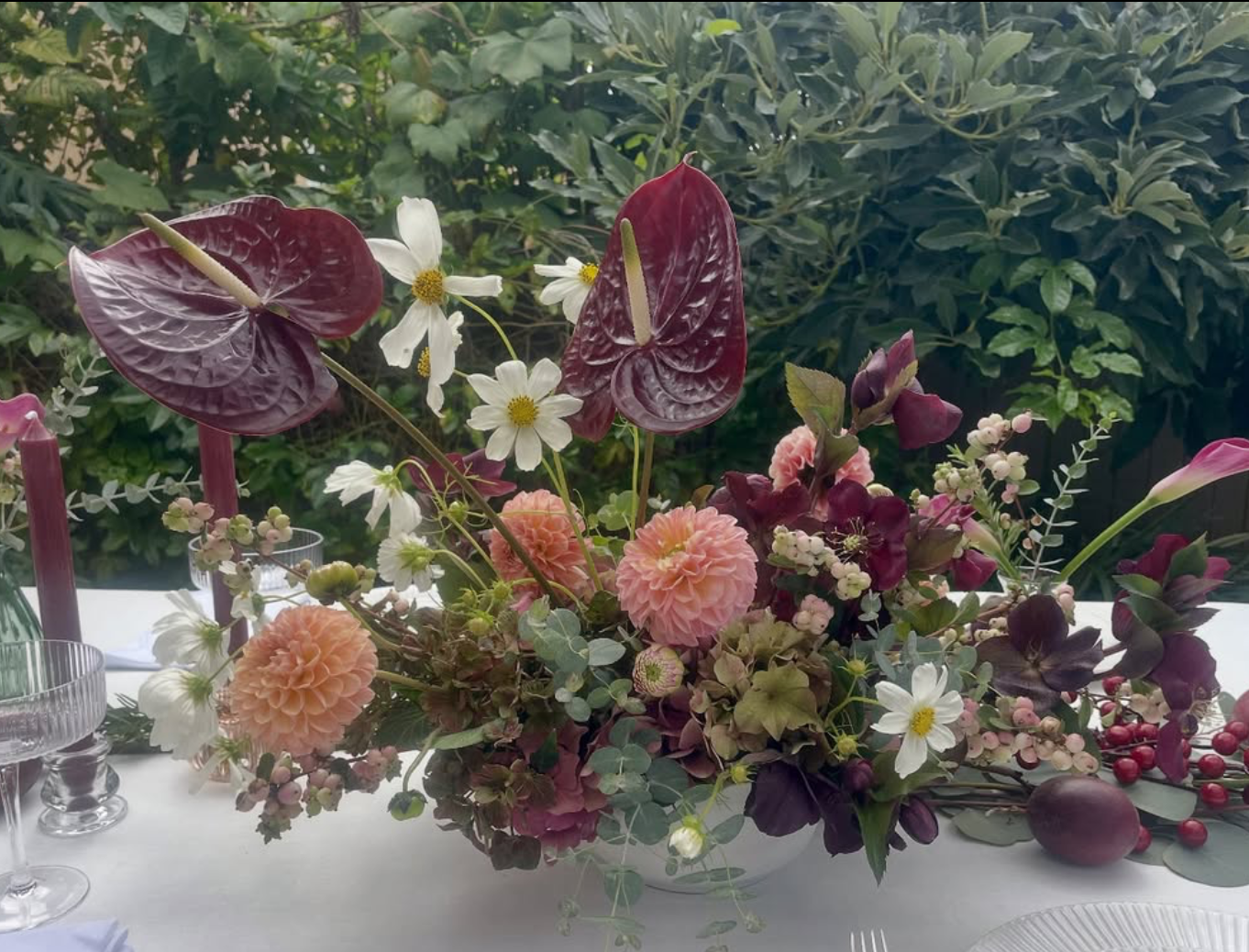 A floral arrangement on a white table with white, pink, and deep purple flowers, including anthuriums, cosmos, dahlias, and berries, against a background of green foliage.