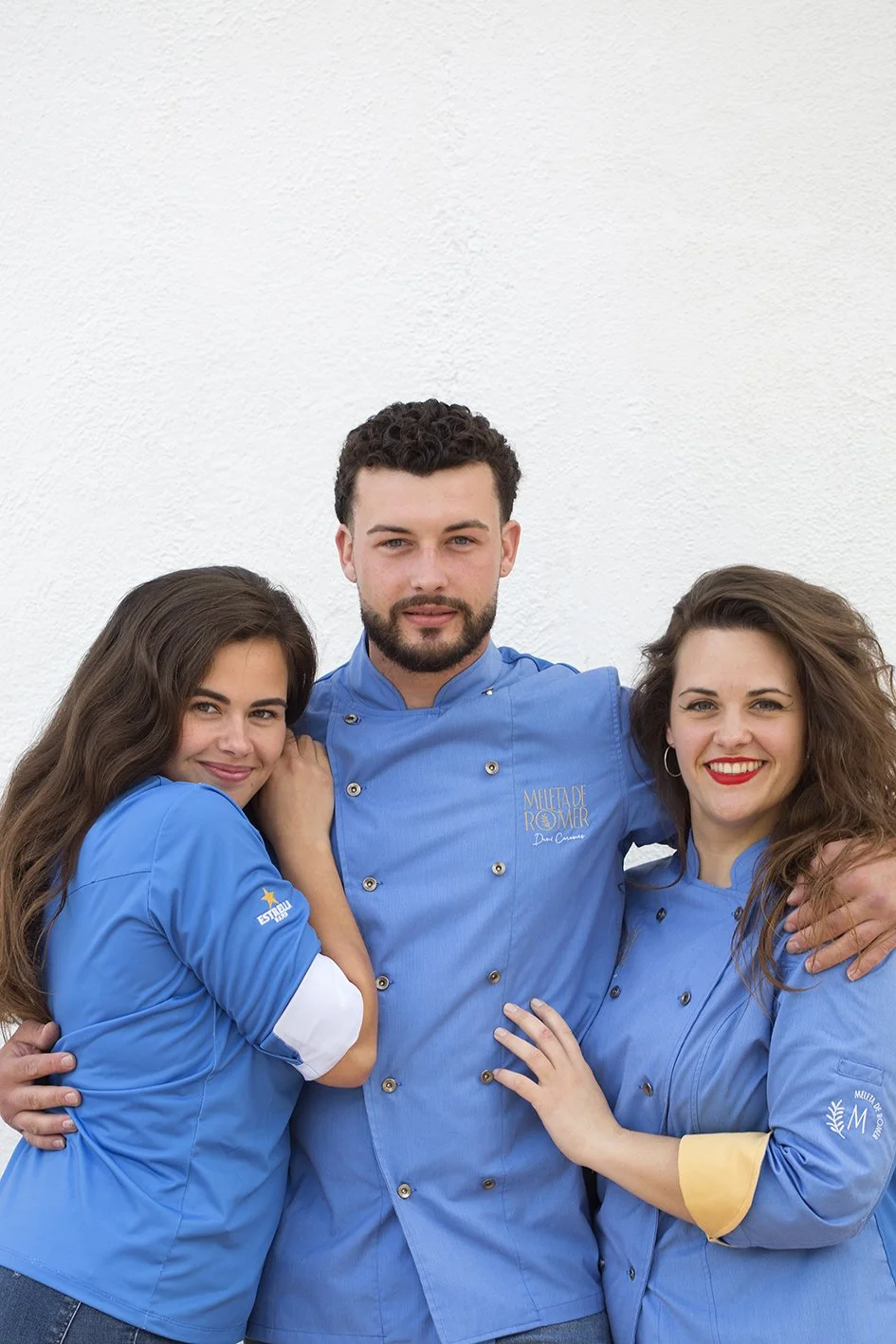 Tres chefs con chaquetas azules, dos mujeres y un hombre, posando juntos frente a una pared blanca.