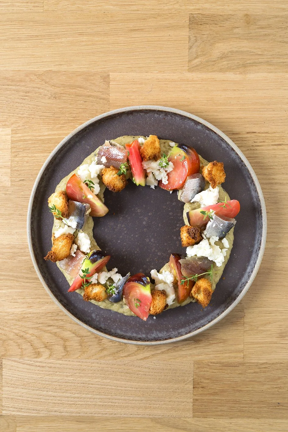 Plato con tomates, queso, sardinas y crutones en un plato oscuro sobre una mesa de madera.