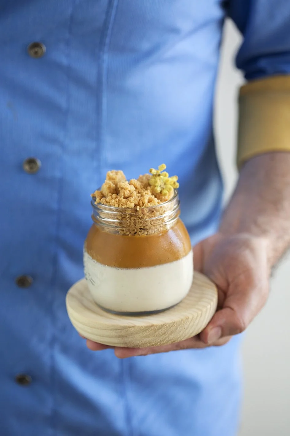 Persona sosteniendo un frasco de postre con capa de caramelo, crema y crumble, sobre un plato de madera.