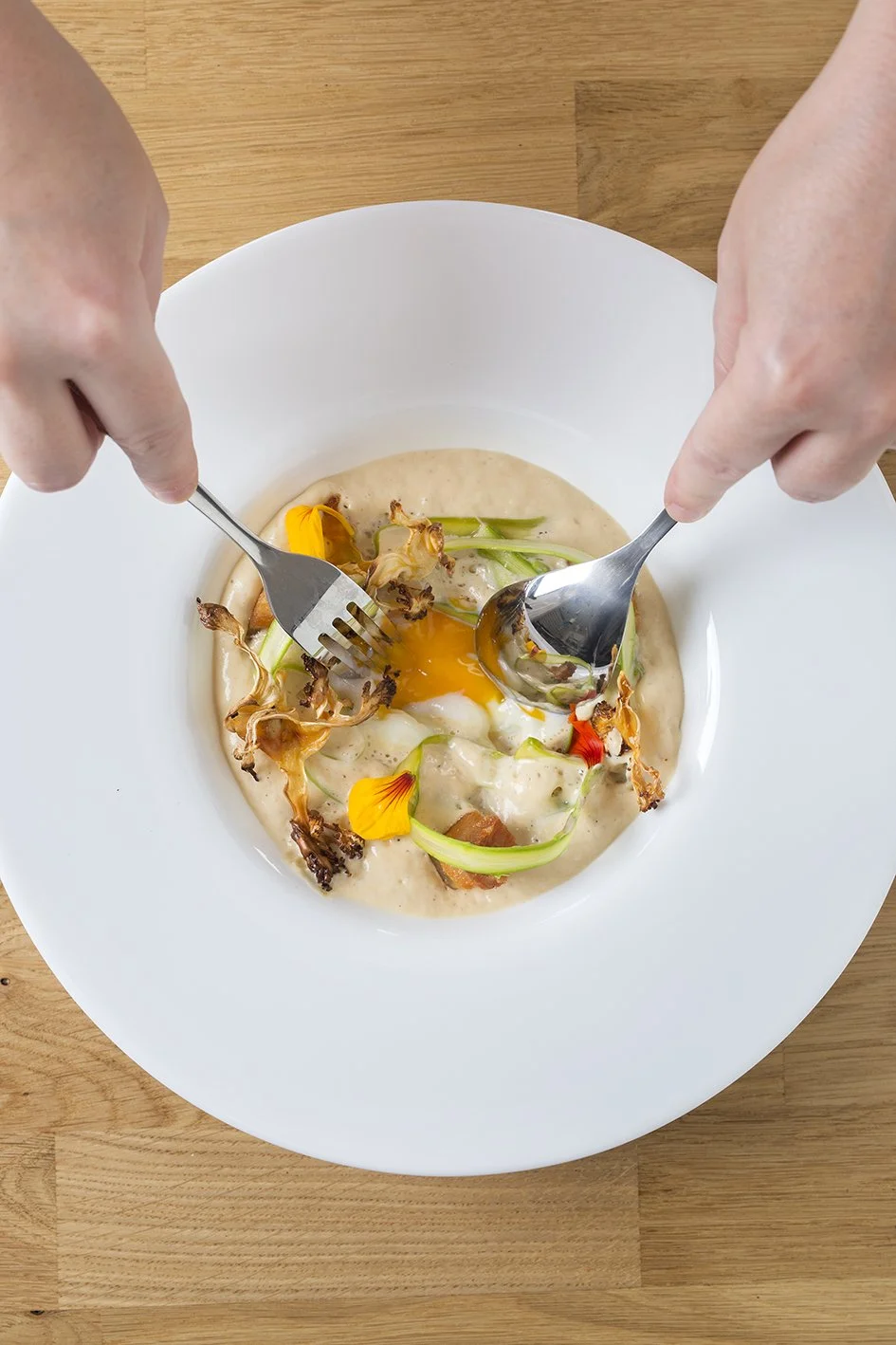 Persona levantando un huevo cocido con tomate, cebolla, y flores comestibles en un plato blanco.