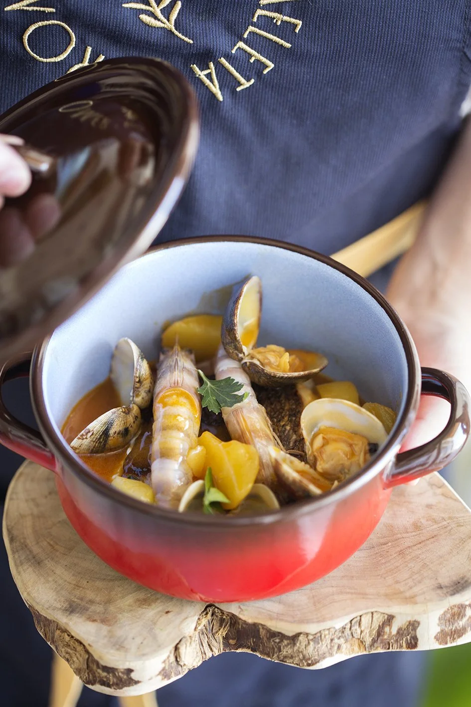 Un plato con mariscos como camarones, almejas y pescado en caldo, servido en una olla roja, con una hoja de perejil encima, sobre una tabla de madera.