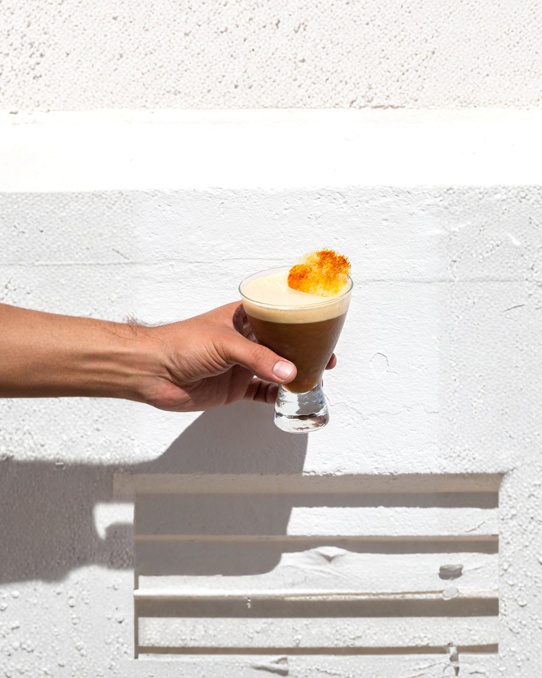 Persona sosteniendo una copa con una bebida fría decorada con una rodaja de fruta, en una pared blanca.