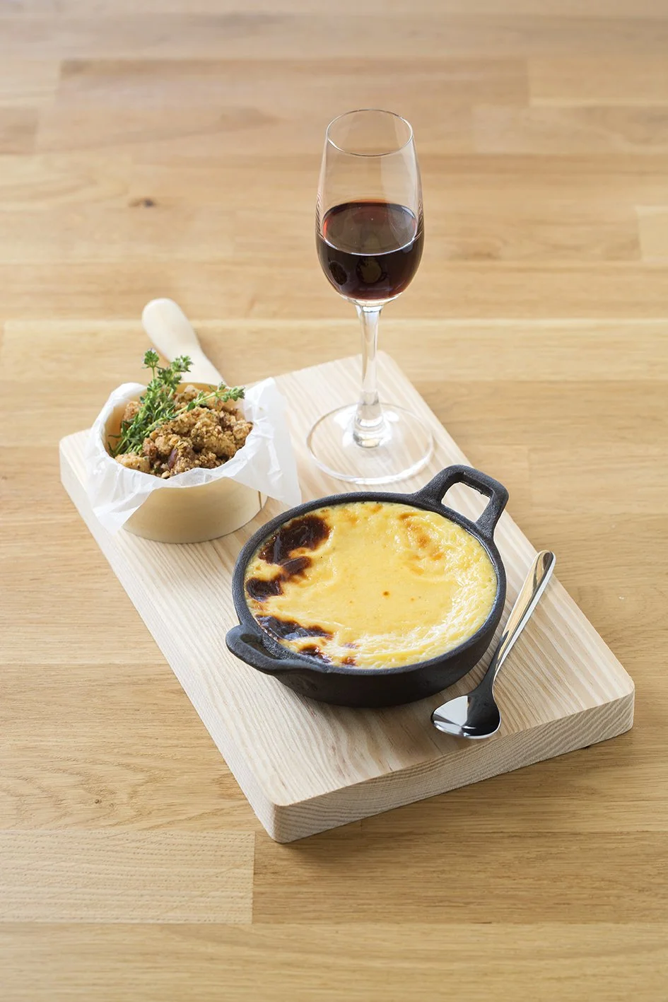 Plato de queso gratinado, tazón con carne y hierbas, copa de vino tinto, todo sobre una tabla de madera.