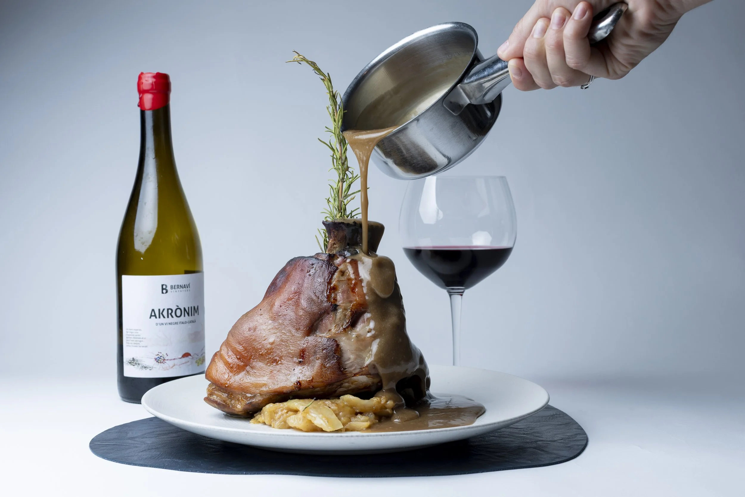 Un plato de carne de res asada con salsa, acompañada de puré de papas y decorada con una ramita de romero, con una copa de vino tinto y una botella de vino a la izquierda.