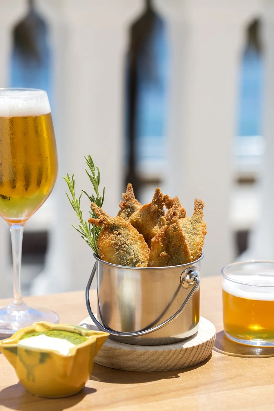 Vaso con cerveza rubia, cuenco con alitas de pollo fritas con romero, bol con salsa y una copa con más cerveza, en una mesa de comedor al aire libre.