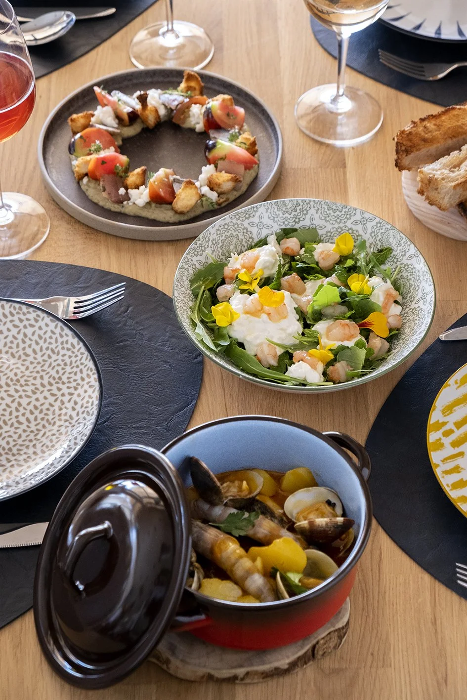Una mesa con varios platos de comida, incluyendo una ensalada con hojas verdes y flores comestibles, una pizza con tomate y pollo, un plato de mejillones en caldo, pan tostado y copas de vino blanco y rosado.