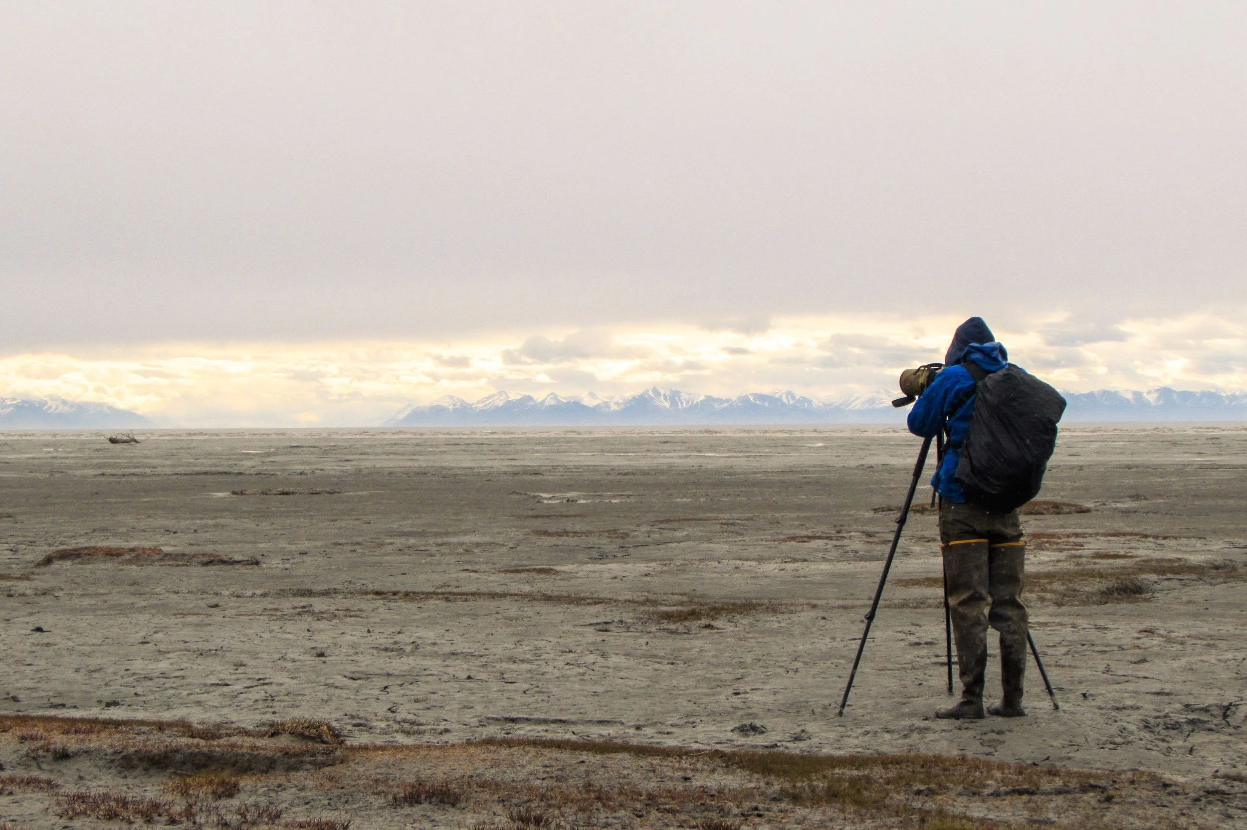 A person in outdoor gear, including a blue jacket, hood, and boots, is standing on a vast, barren landscape with a camera on a tripod, aiming at snow-capped mountains in the distance under a cloudy sky.