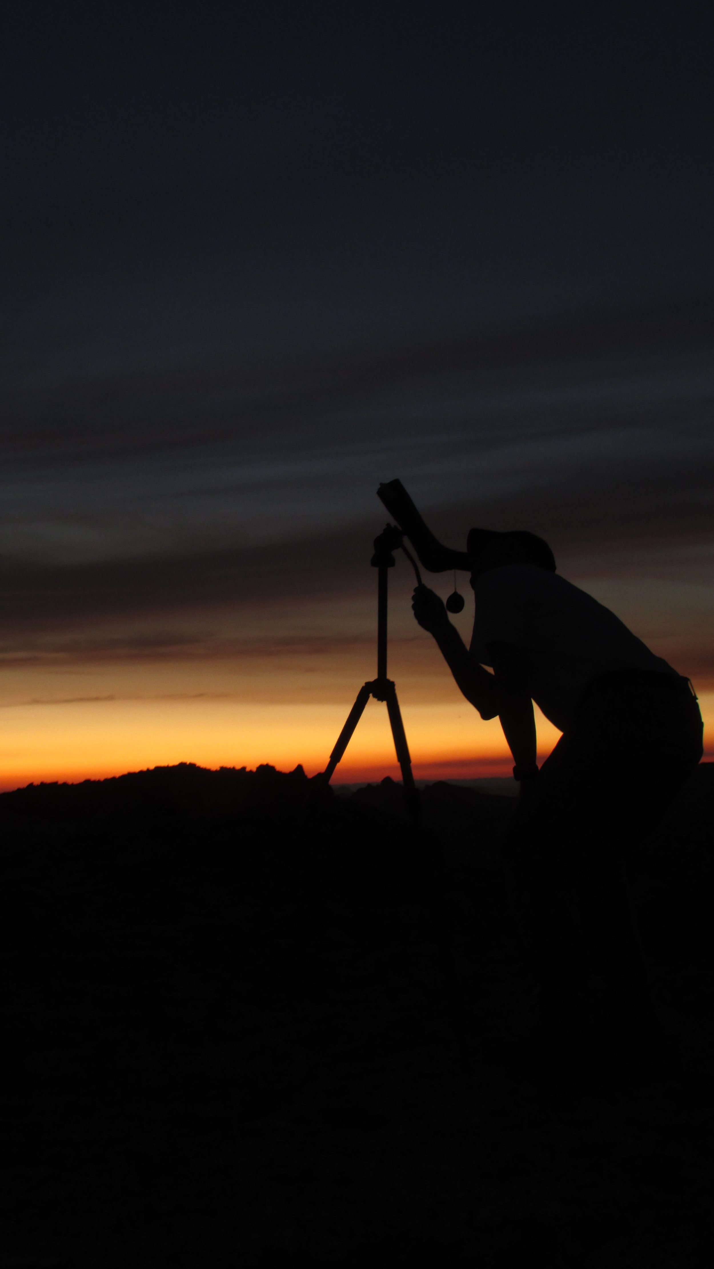 Silhouette of a person looking through a telescope at sunset with a dark sky and orange horizon.