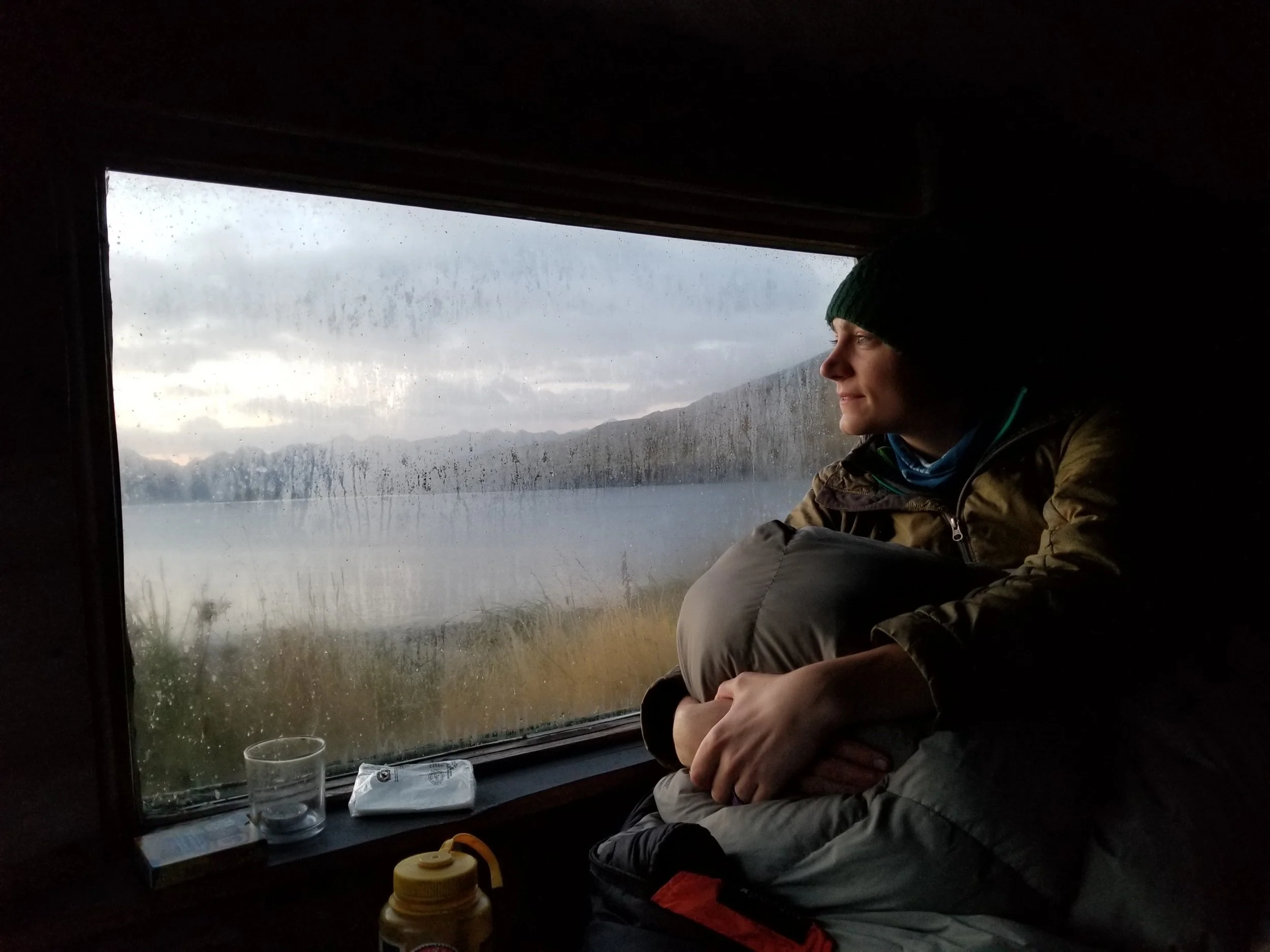 Woman in a beanie hat sits inside a cabin, looking out the window at a misty lake and distant mountains.