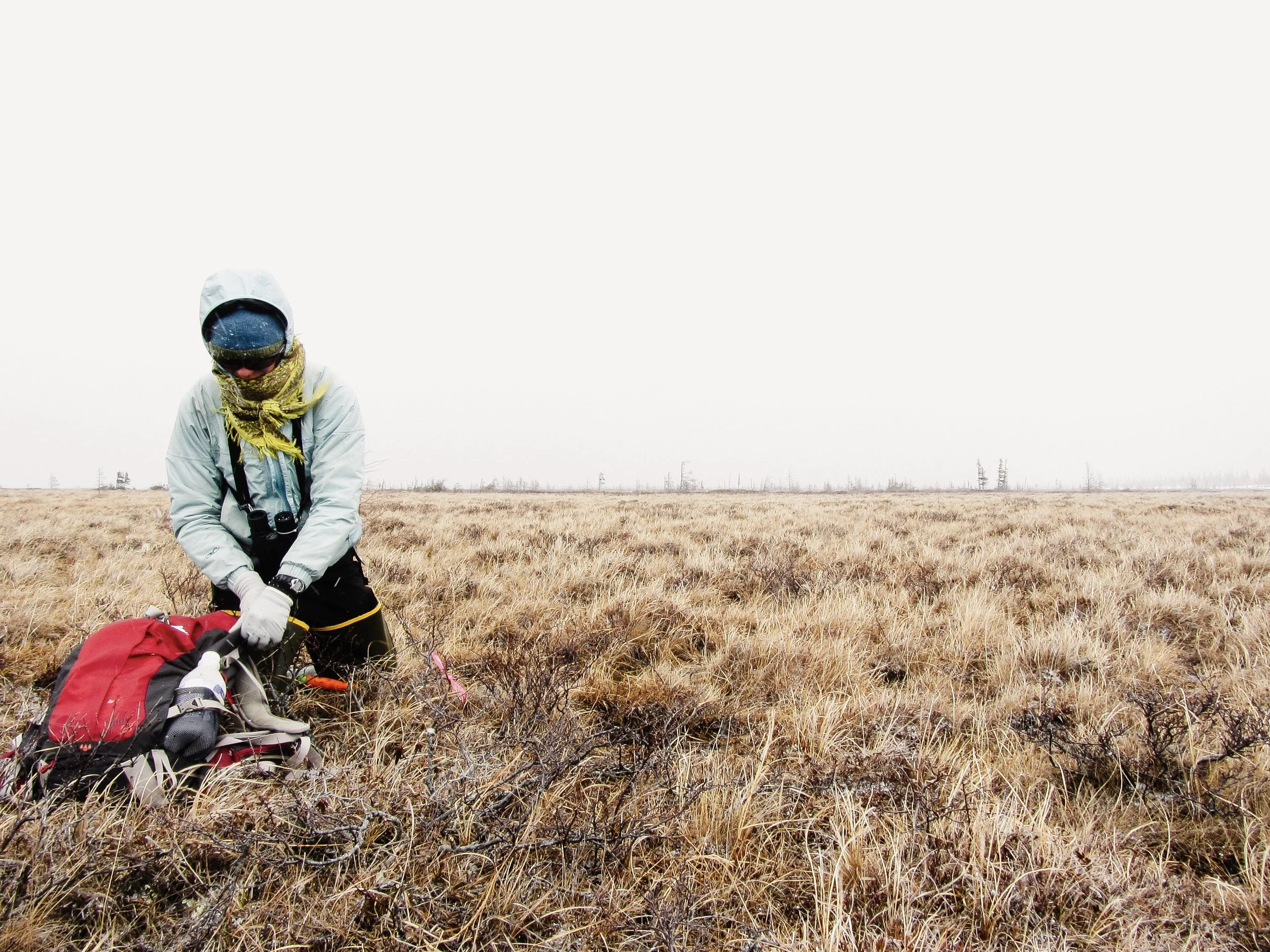 A person wearing outdoor gear, including a hoodie, scarf, goggles, and gloves, stands in a dry, grassy field with a red backpack, appearing to prepare or organize items.