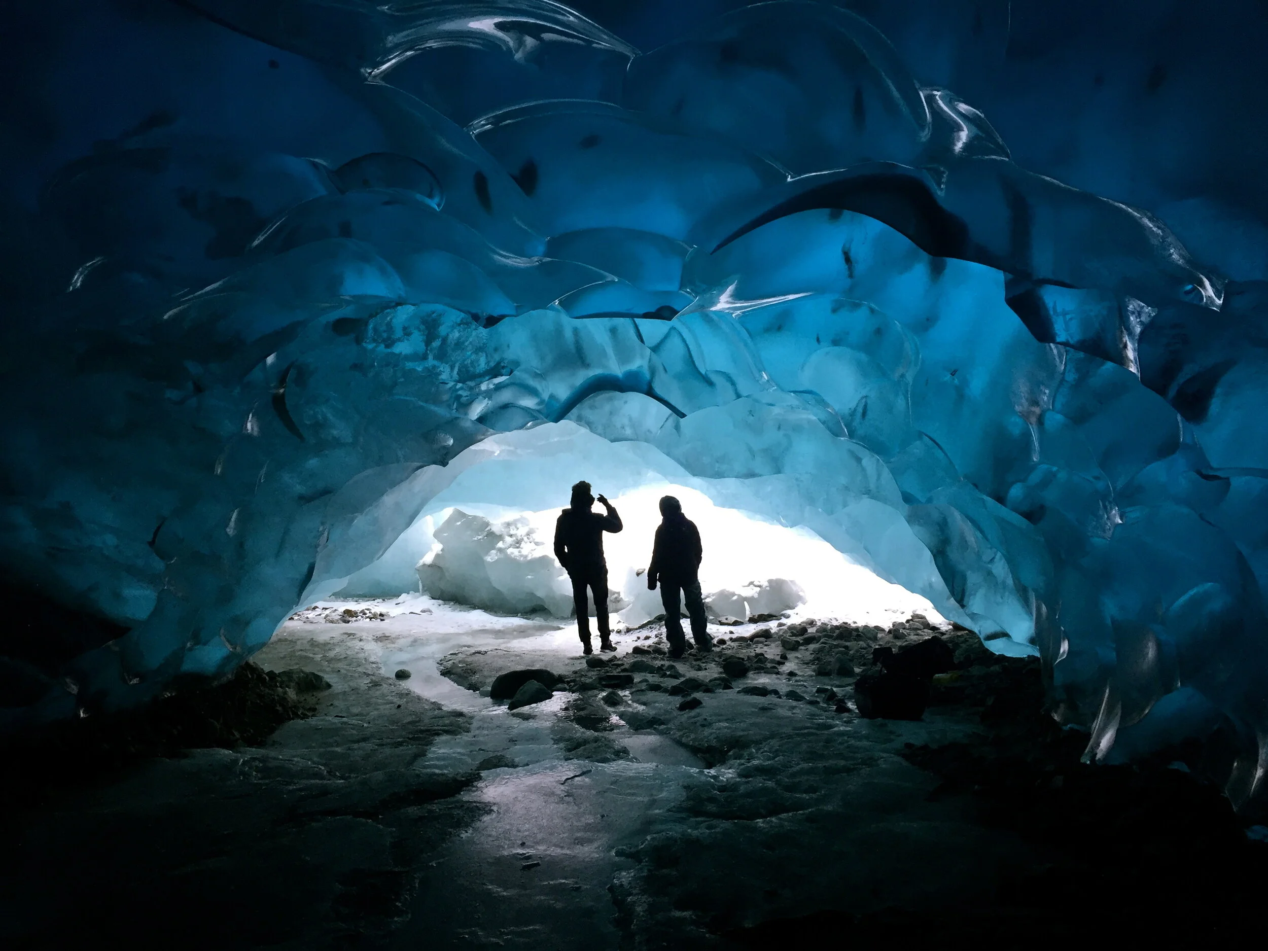 Two people standing inside an ice cave with blue ice formations overhead and sunlight shining through the cave opening.