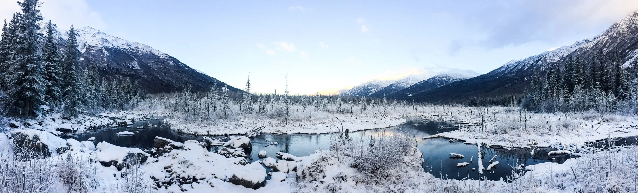 Snow-covered river flowing through a valley with evergreen trees and snow-capped mountains in the background, under a partly cloudy sky.