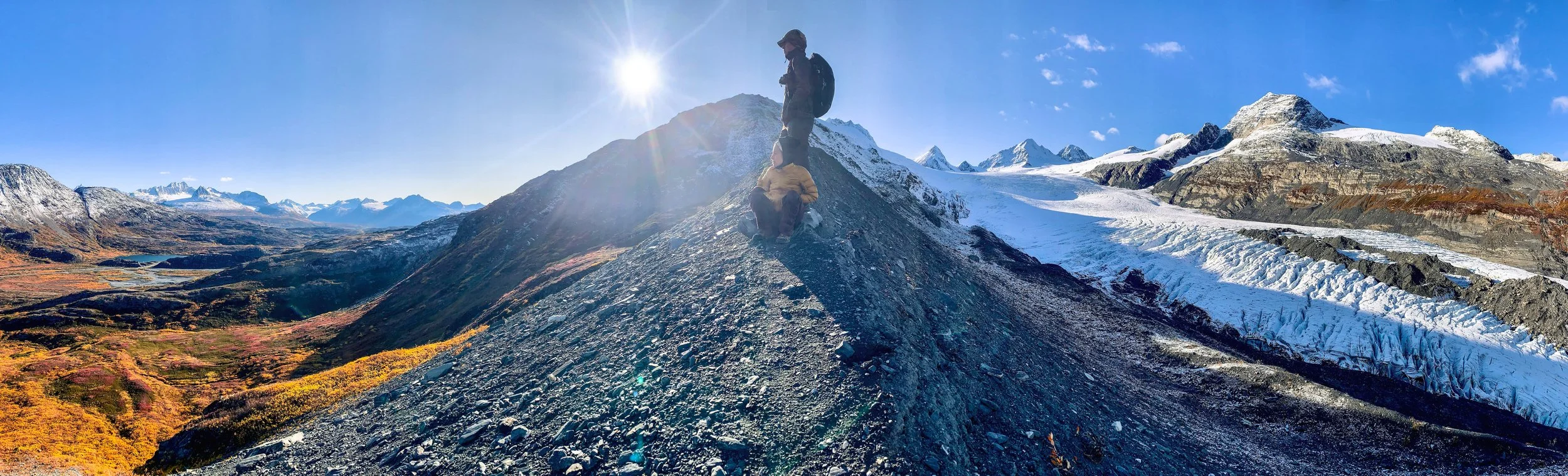 Two hikers, one standing and one sitting, on a rocky mountain ridge with snow-covered peaks in the background under a clear blue sky and bright sun.