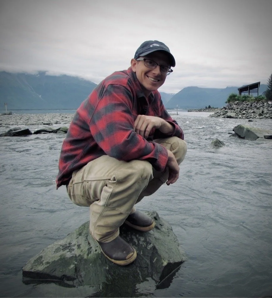 A man wearing glasses, a dark cap, a red and black checkered flannel shirt, khaki pants, and Xtratuf boots squats on a rock in an estuary, smiling at the camera with mountains and cloudy skies in the background.