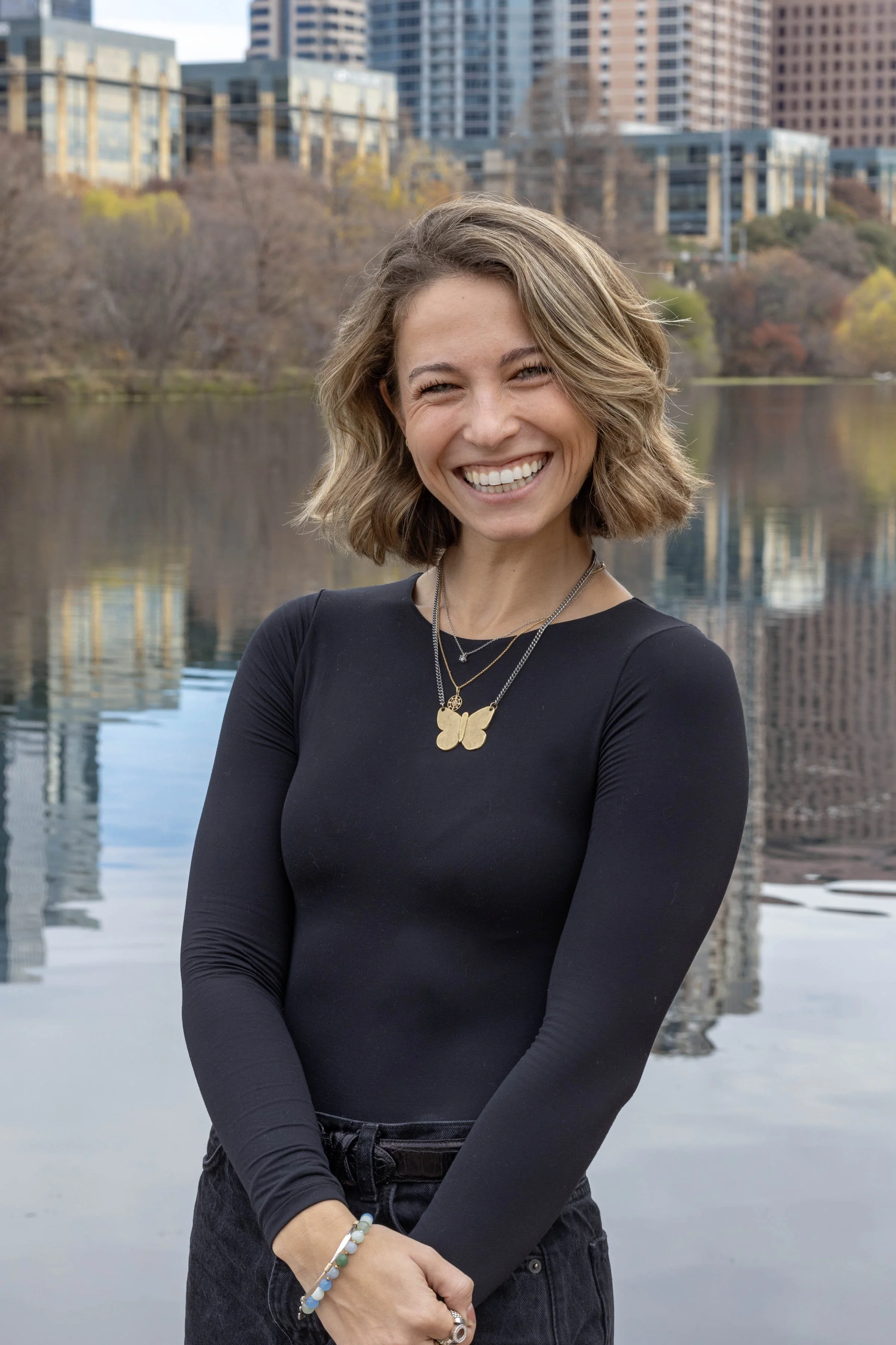 A woman with shoulder-length wavy hair smiling outdoors by a body of water with city buildings in the background.