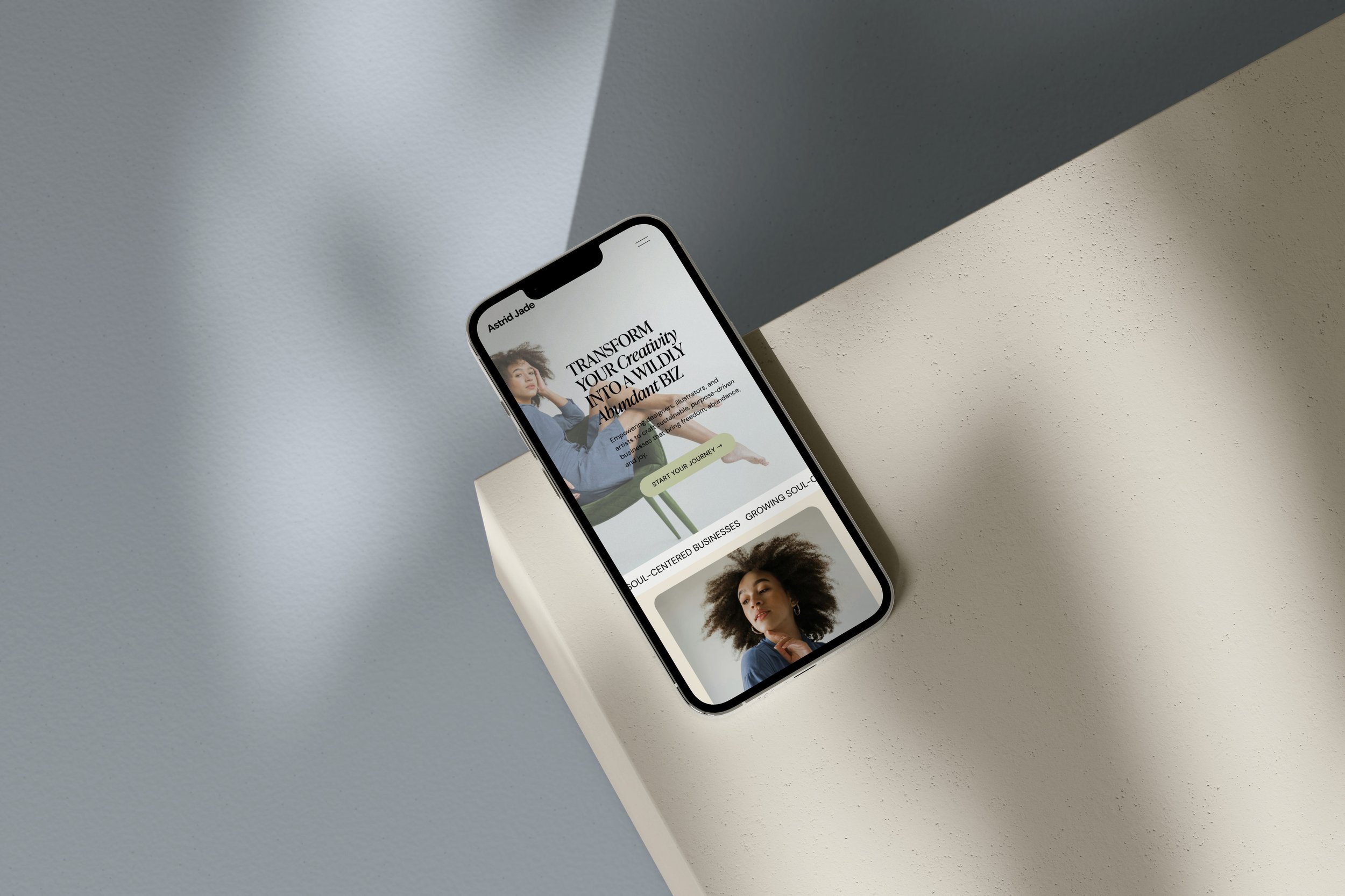 An iPhone lying on a white pedestal, displaying a mobile-friendly website built with the drag-and-drop Astrid Jane website template for Squarespace from Heythere Studio.