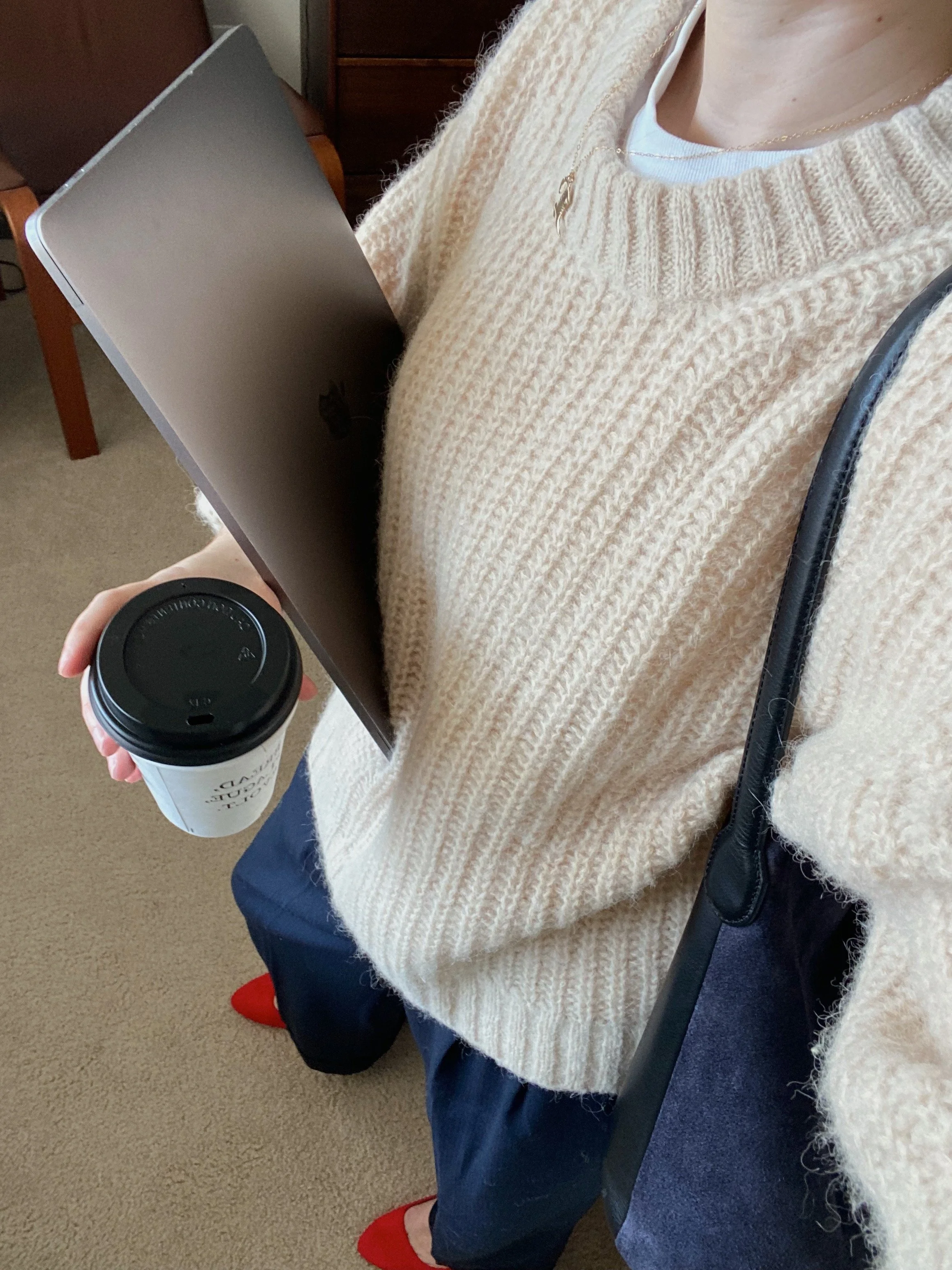 Person wearing a beige sweater holding a laptop and a cup of coffee, with a black strap purse over shoulder, standing on a carpeted floor.