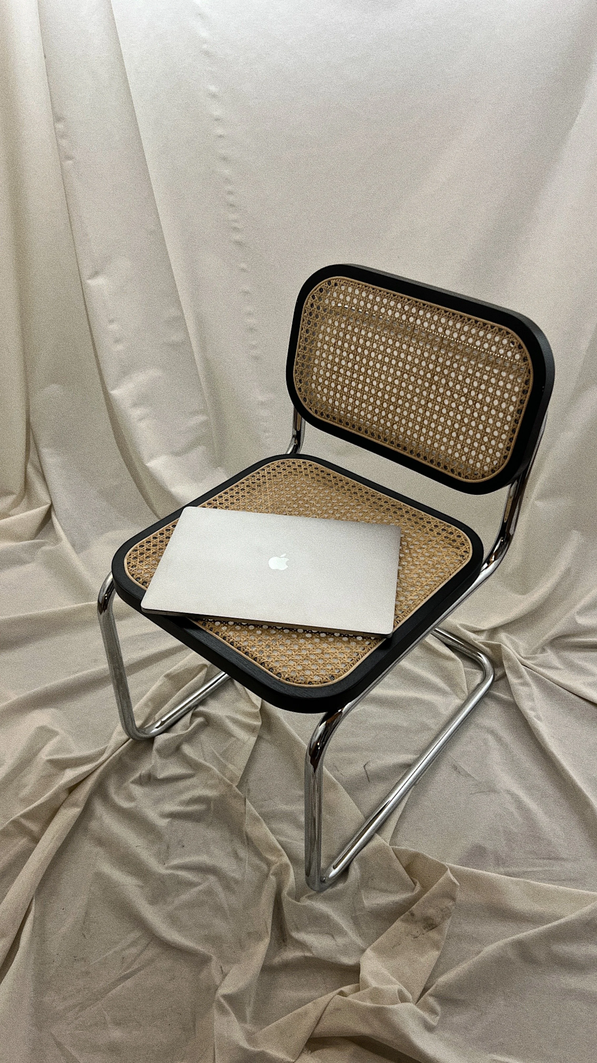 A chair with a woven cane seat and backrest, and a silver metal frame, with a closed silver MacBook laptop on the seat. Beige fabric drapes in the background and on the floor.
