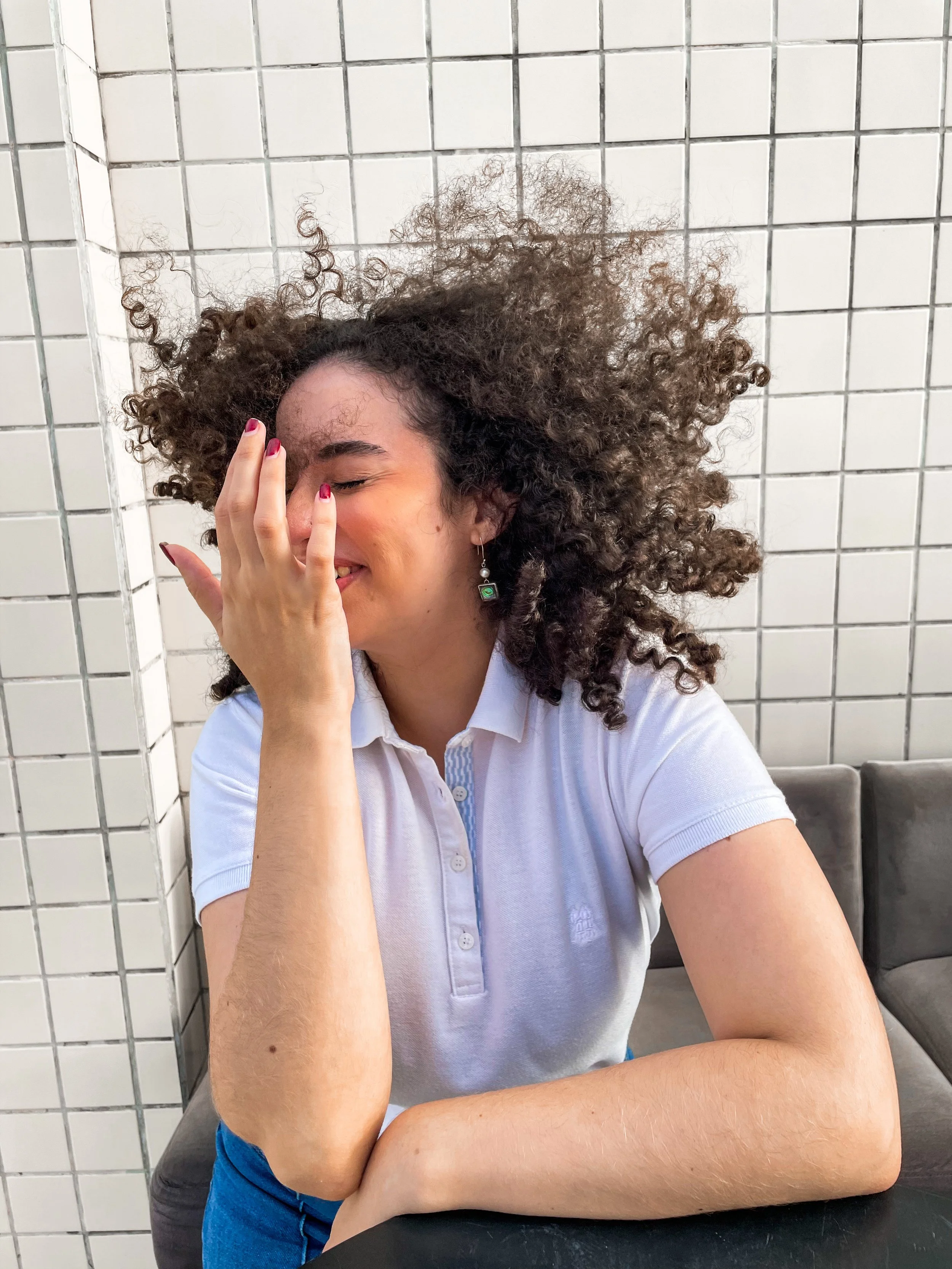 A woman with curly hair wearing a white polo shirt, sitting with her right hand covering her face, smiling.