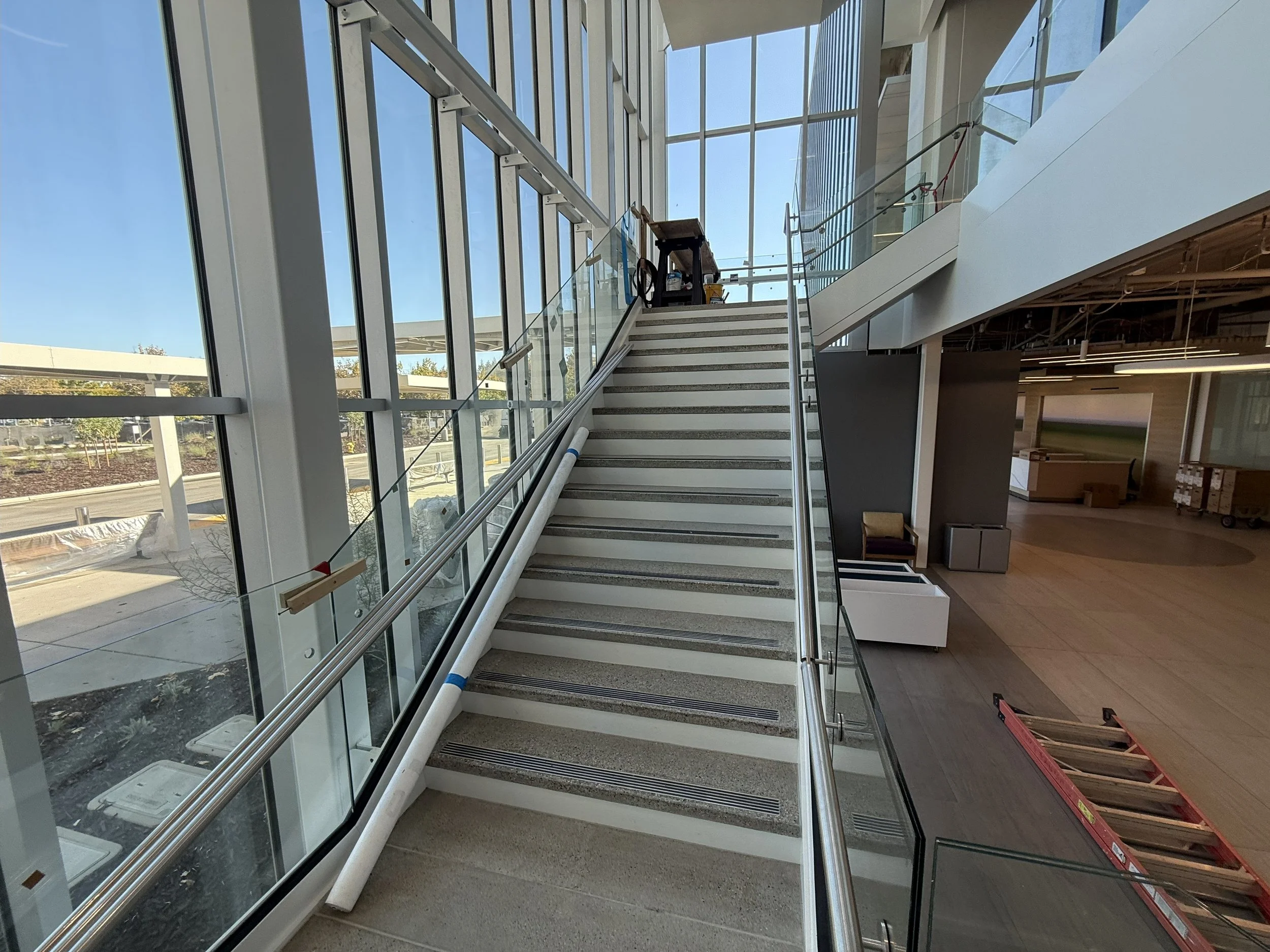 Interior view of a building staircase under construction, with construction materials and tools around, and large glass windows letting in natural light.