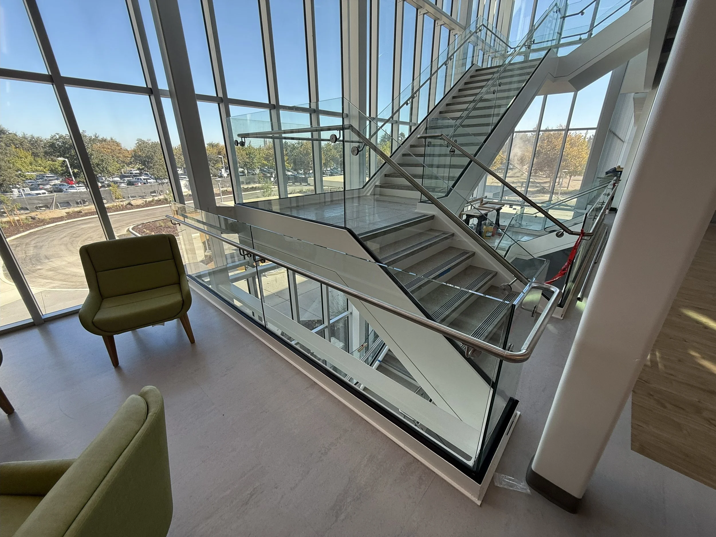 Interior view of a modern building with large glass windows, showing staircase with glass railings and a seating area with green chairs.