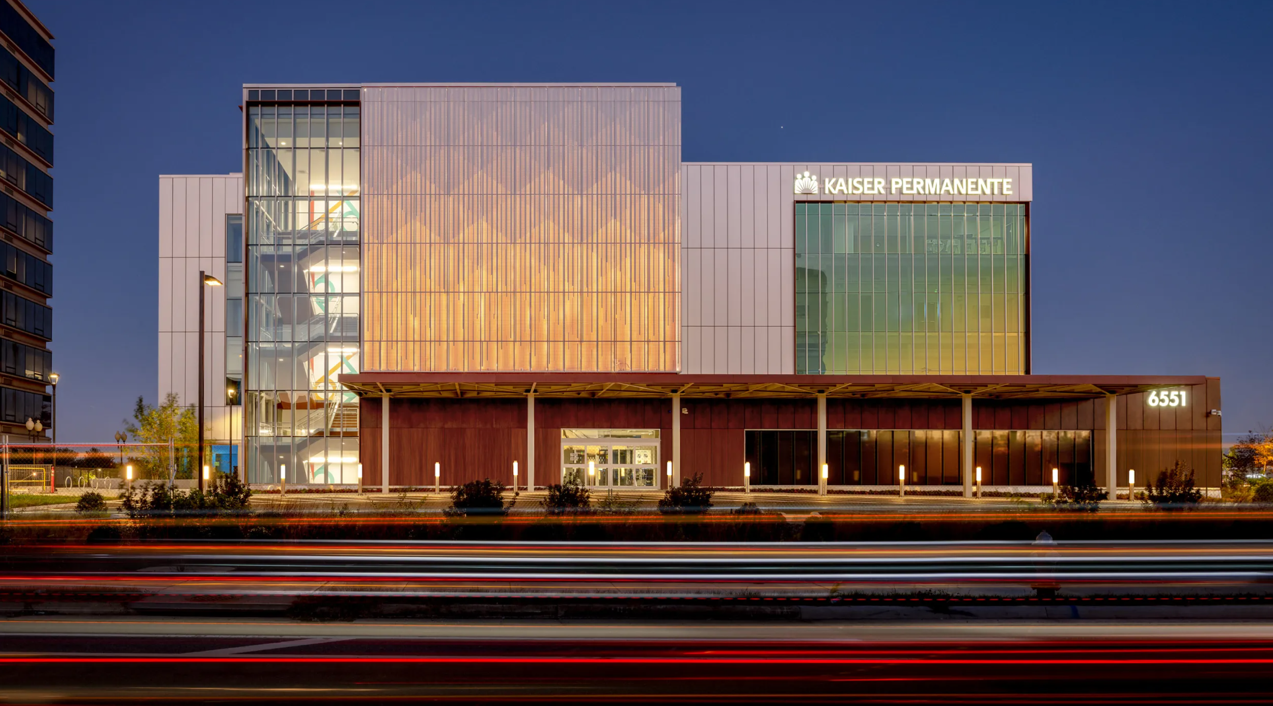 Modern hospital building with 'Kaiser Permanente' sign, glass windows, and a visible spiral staircase inside, at dusk with light trails from passing cars in front.