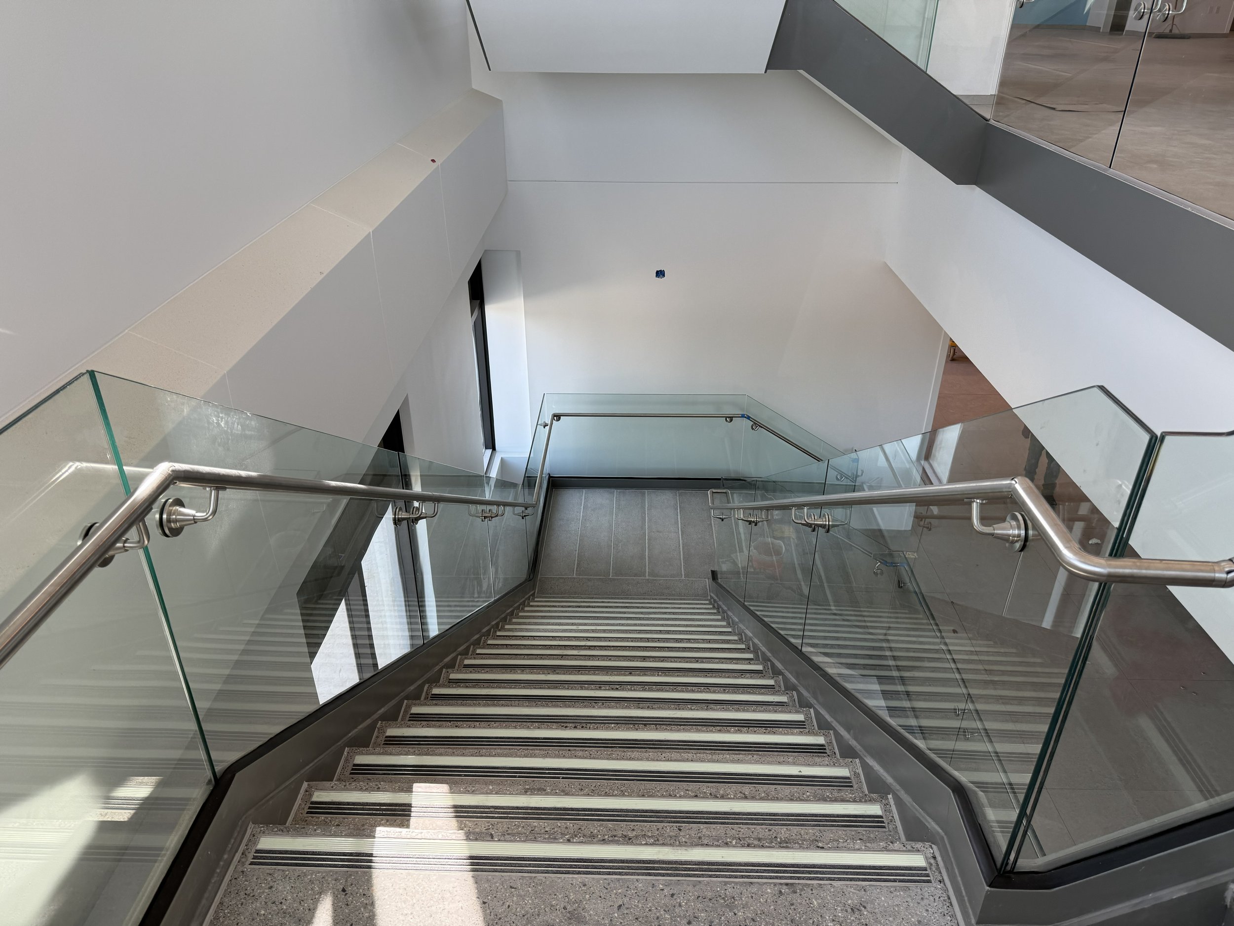 A downward view of a modern stairway with marble steps, glass railings, and metal handrails inside a building with white walls.