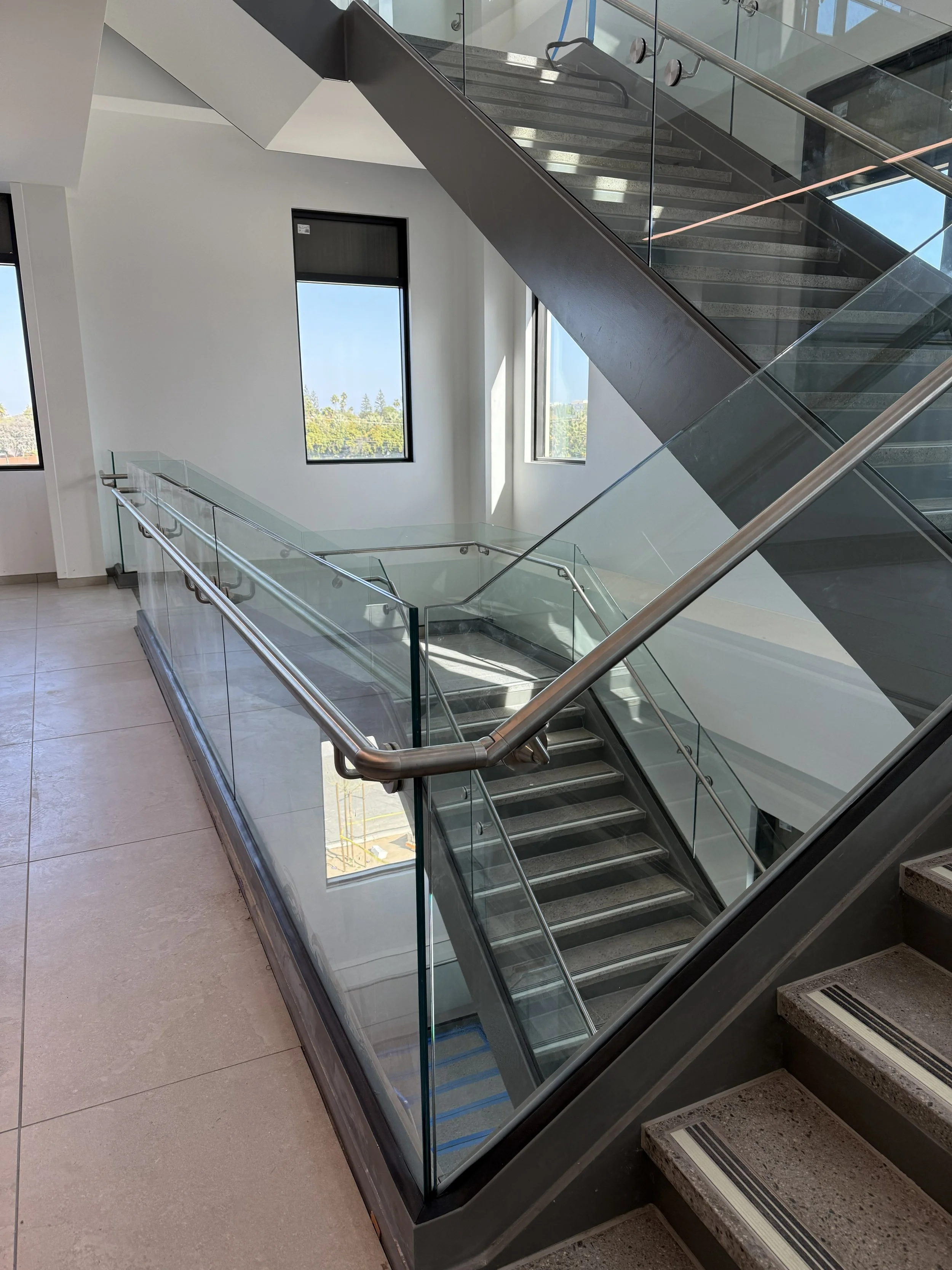 Modern staircase inside a building with glass railings, metal handrails, and large windows providing natural light.