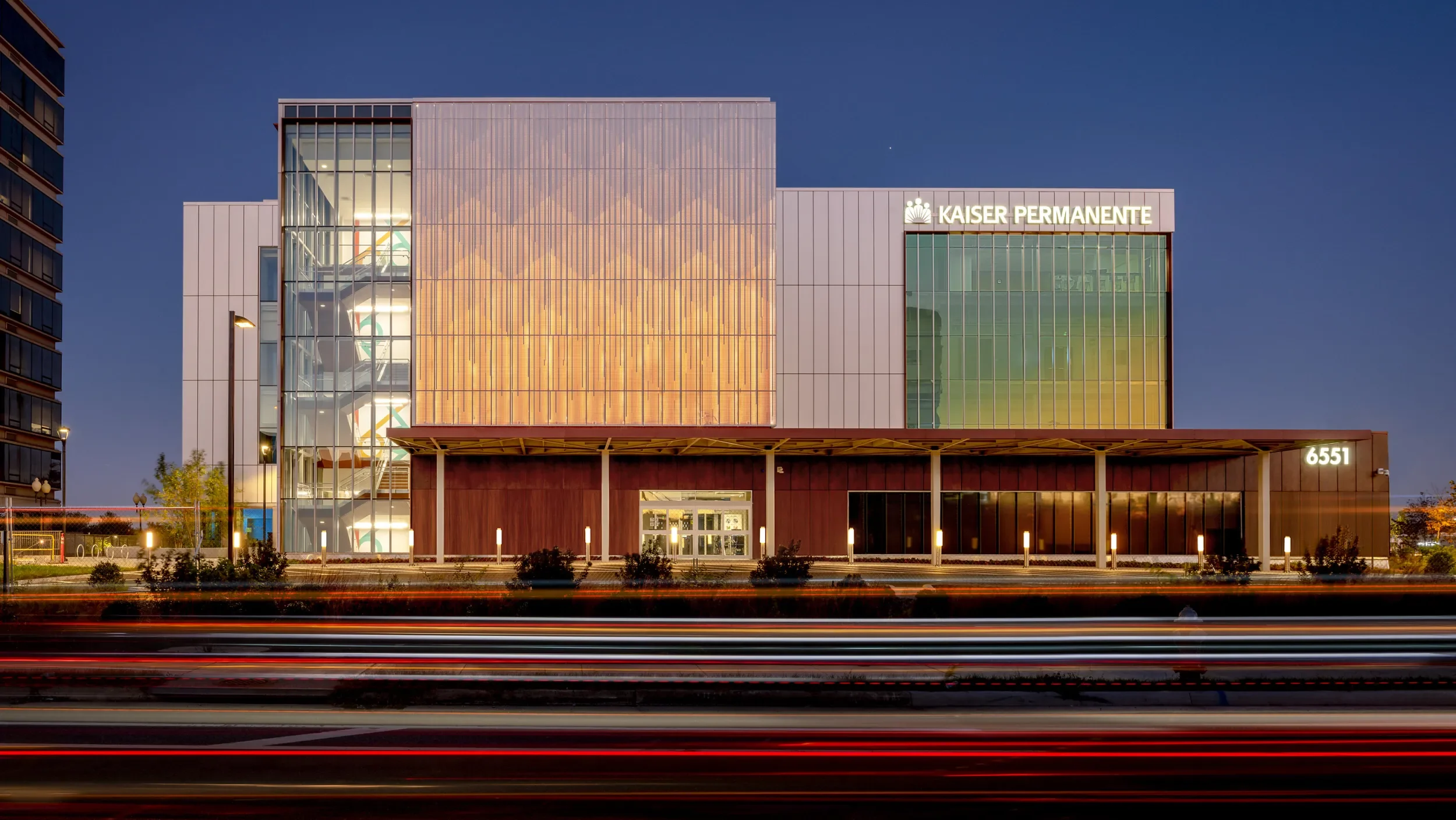 Exterior of a modern hospital building at night with the sign 'Kaiser Permanente' and the address '6551', illuminated and with streaks of car lights in the foreground.