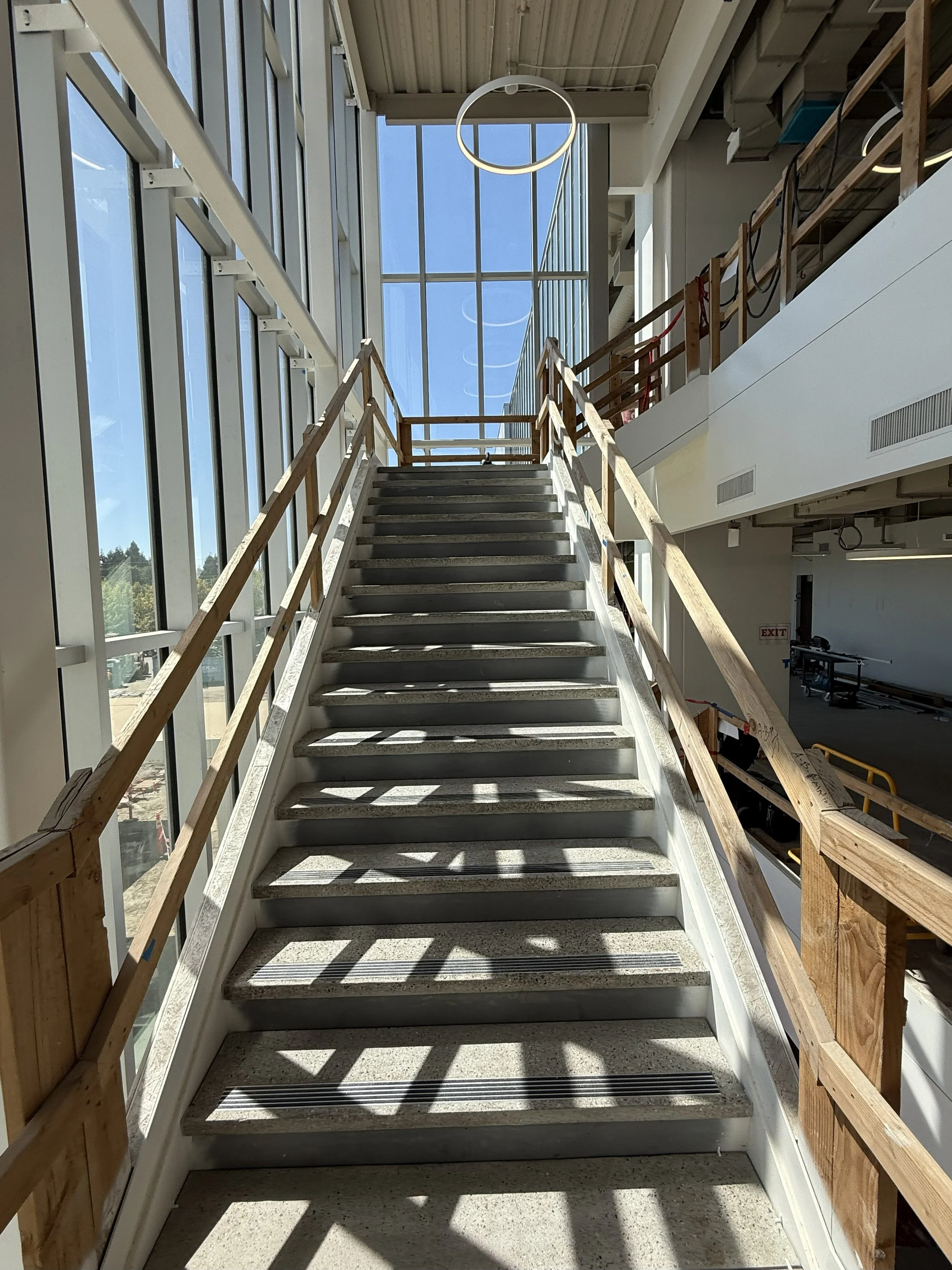 Indoor construction site with a staircase leading upward, surrounded by large glass windows allowing natural light, and partially installed wooden railings.