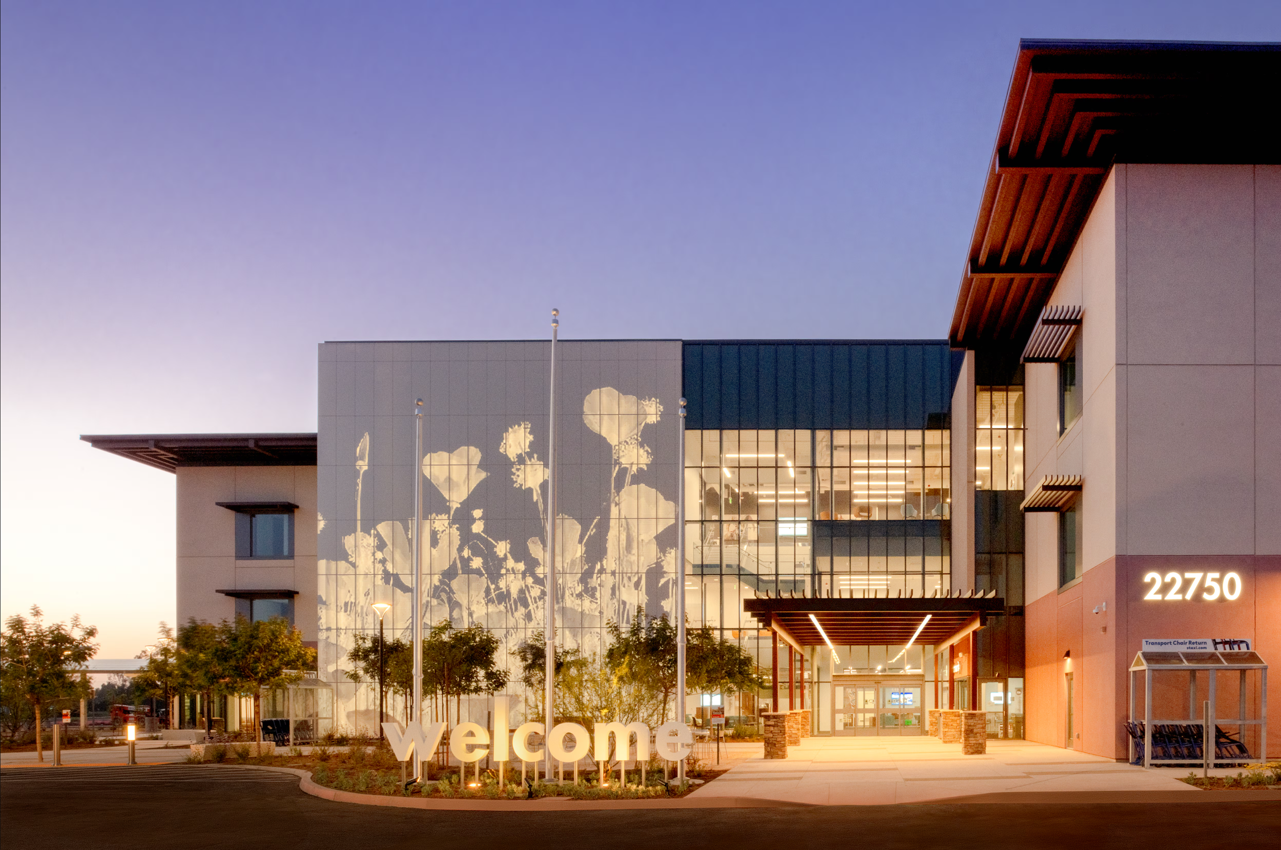 Modern building with large glass windows, trees, and a welcome sign in front, illuminated at dusk.