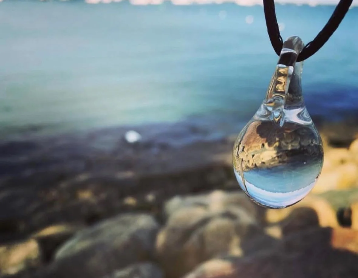 A glass teardrop-shaped pendant with an image of a rocky shoreline and water inside, hanging from a black cord, outdoors near a rocky beach with water in the background.