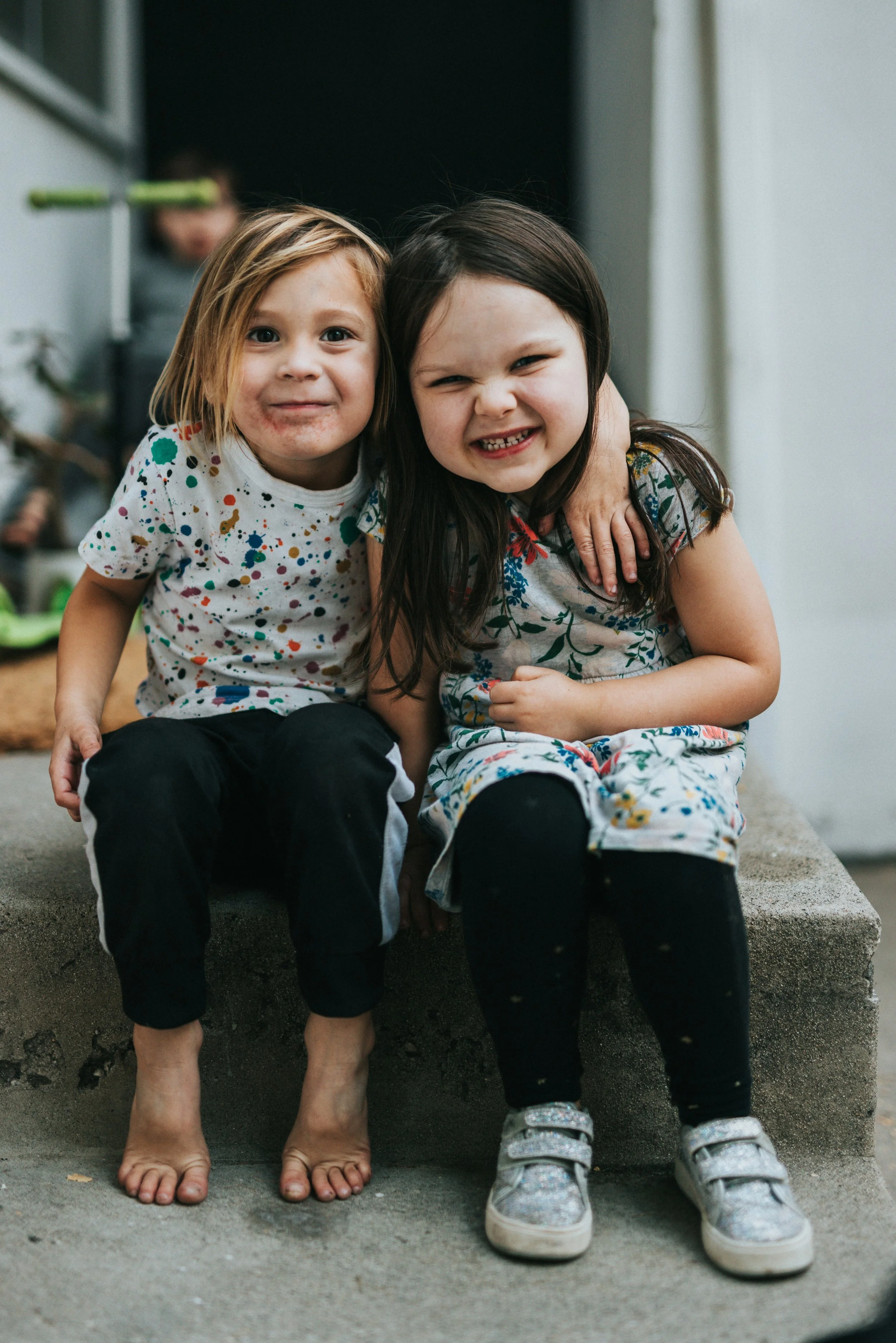 Two young girls sit closely together on a concrete step, smiling and showing a friendly and playful bond. One girl has light skin and blonde hair, wearing a multicolored splattered shirt and black pants, while the other has light skin and dark hair, wearing a floral dress and shiny sneakers. Behind them, a boy is slightly blurred in the background.